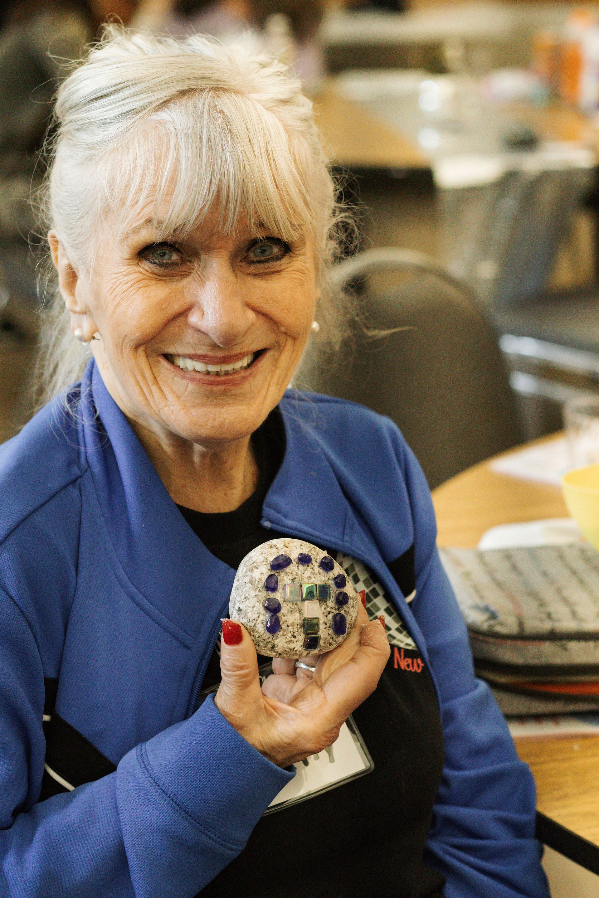 Woman with white hair smiling, holding a decorative mosaic-covered object, indoors. She wears a blue jacket.
