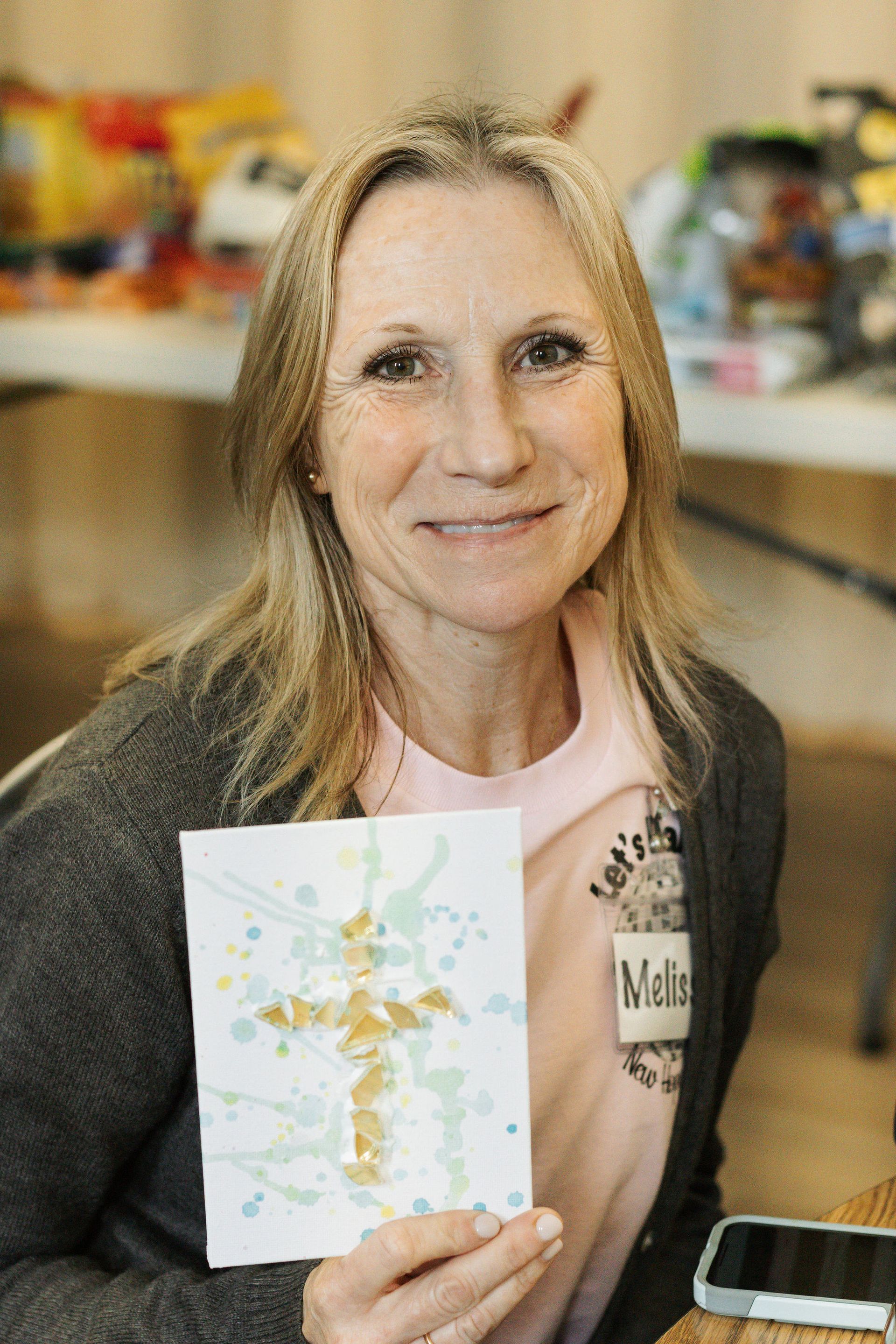 Woman smiling, holding a watercolor cross painting, with blonde hair, gray cardigan, and light pink shirt.