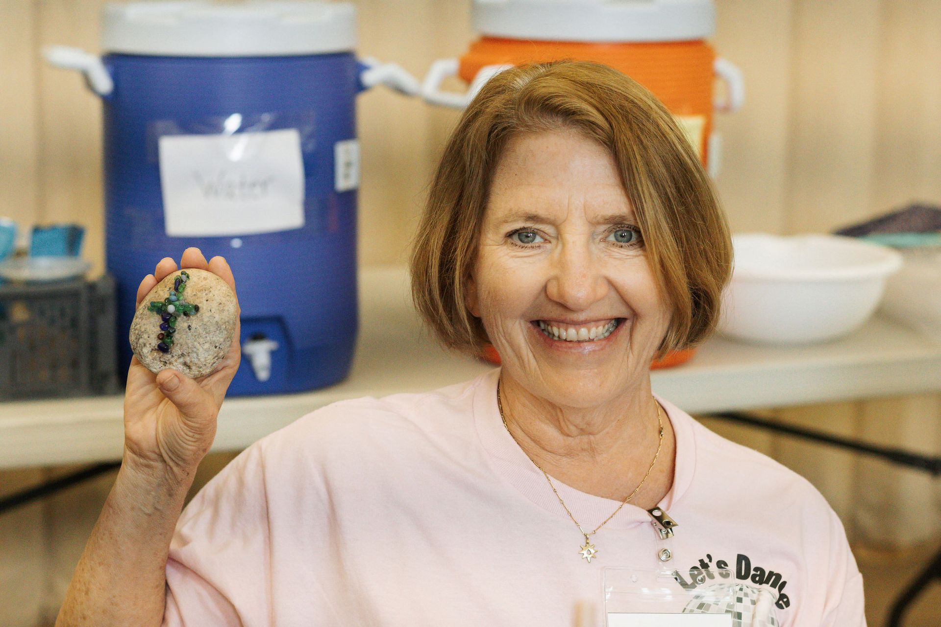 Woman smiling, holding painted rock with a cross; water cooler in background.