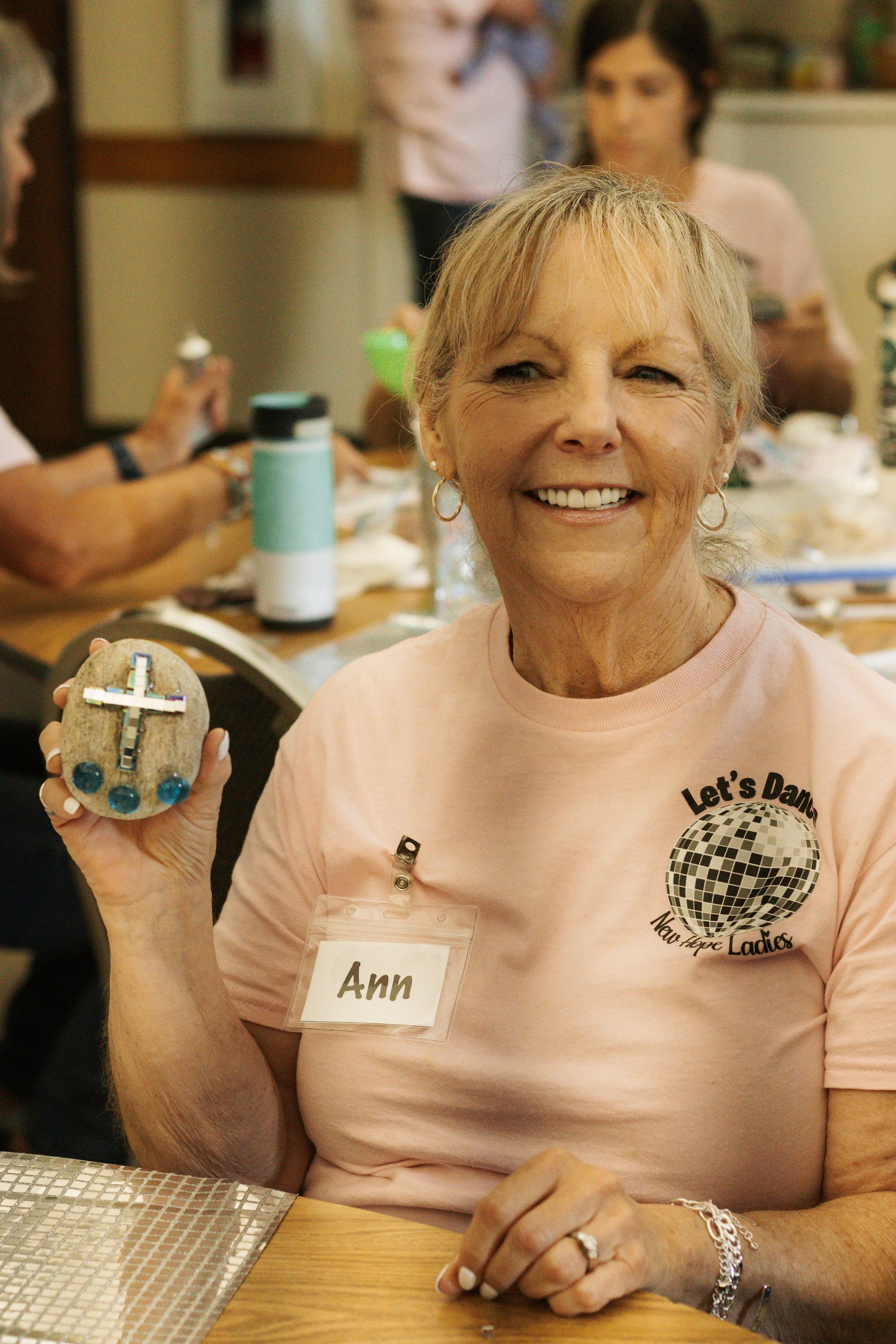 Smiling woman named Ann holds a rock decorated with a cross, likely at a church event.