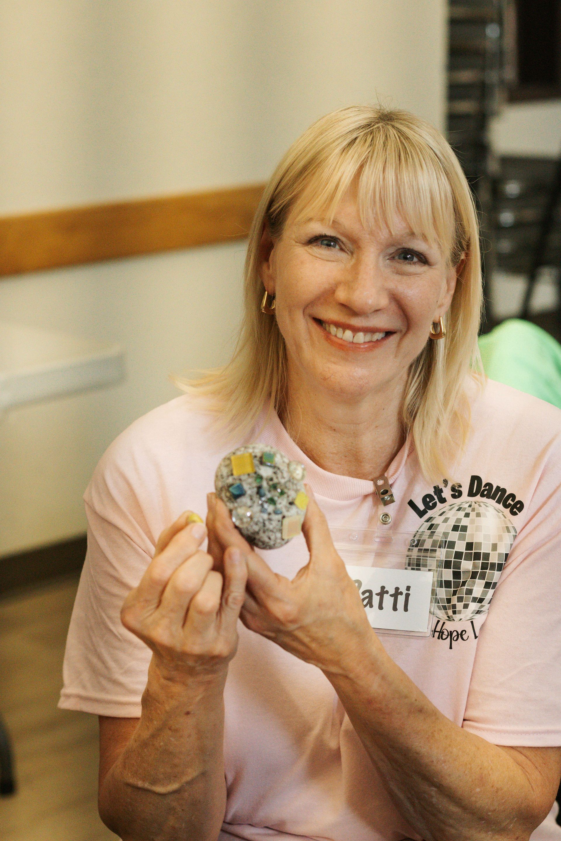 Woman smiles, holding a colorful stone. She wears a pink shirt indoors.
