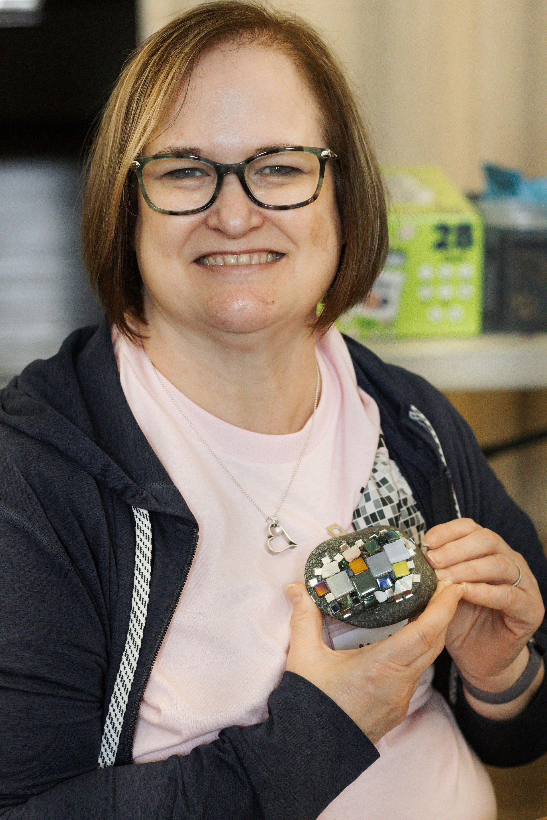 Woman with glasses, smiling, holds a mosaic craft with multiple colors.