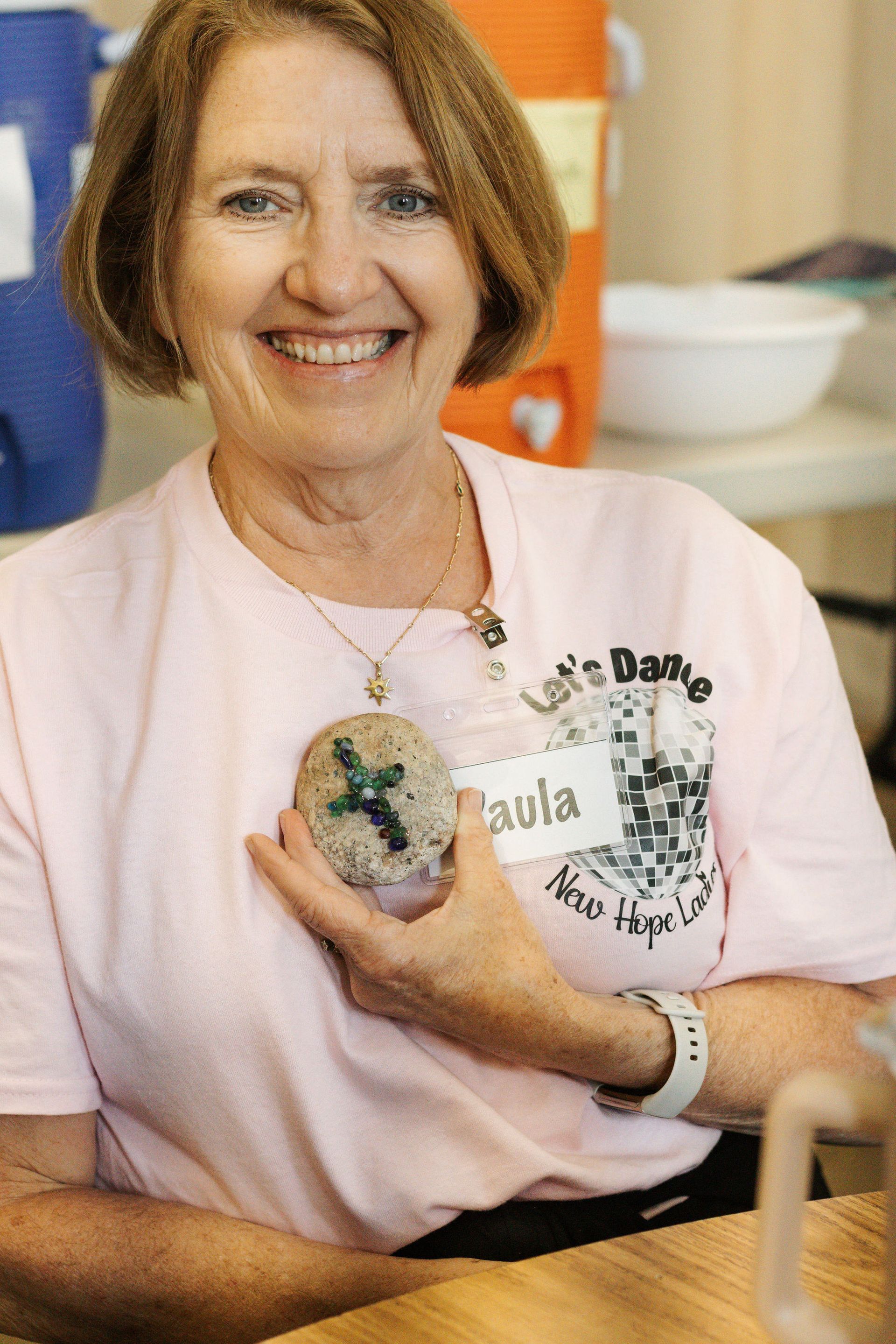 Woman named Paula smiles, holding a decorated stone, wearing a pink shirt that says 