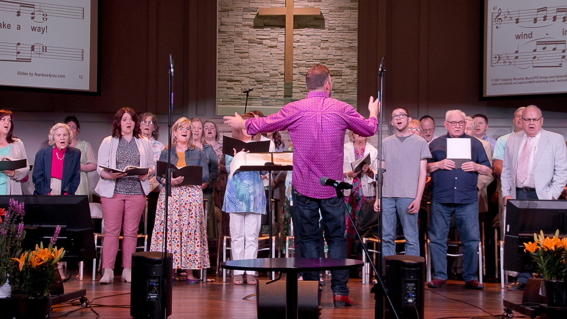 A man in a purple shirt is leading a choir on a stage.