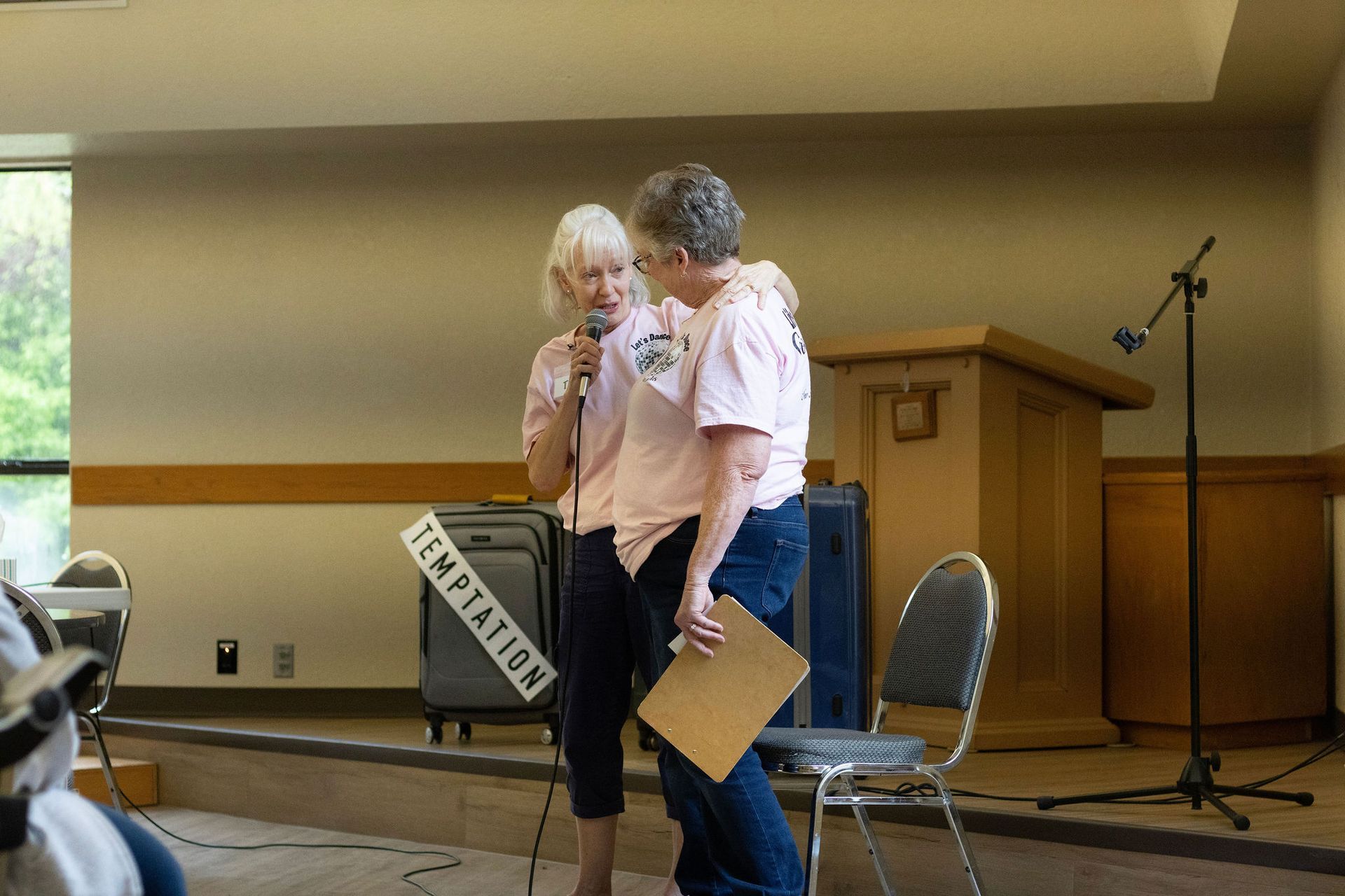 Two women, hugging, wearing pink shirts, one holding a microphone, speaking on a stage, 