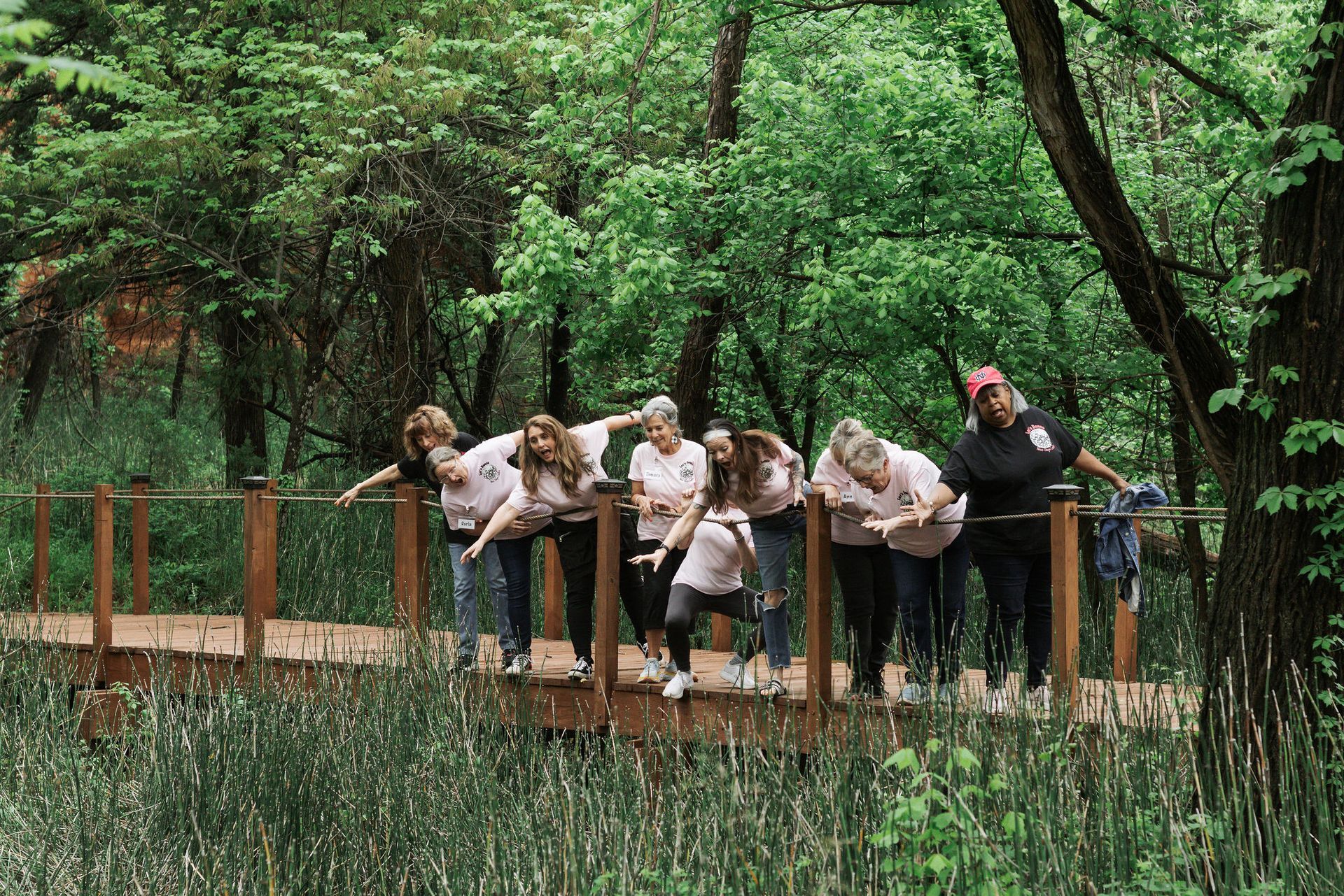 Group of people leaning over a wooden bridge in a green forest; some wearing pink shirts.