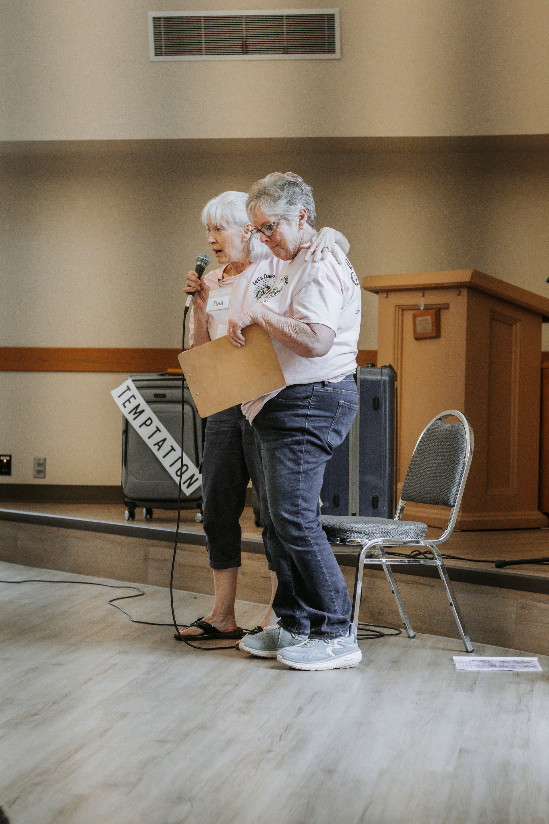 Two older women on stage, one with a microphone, holding a sign that says 