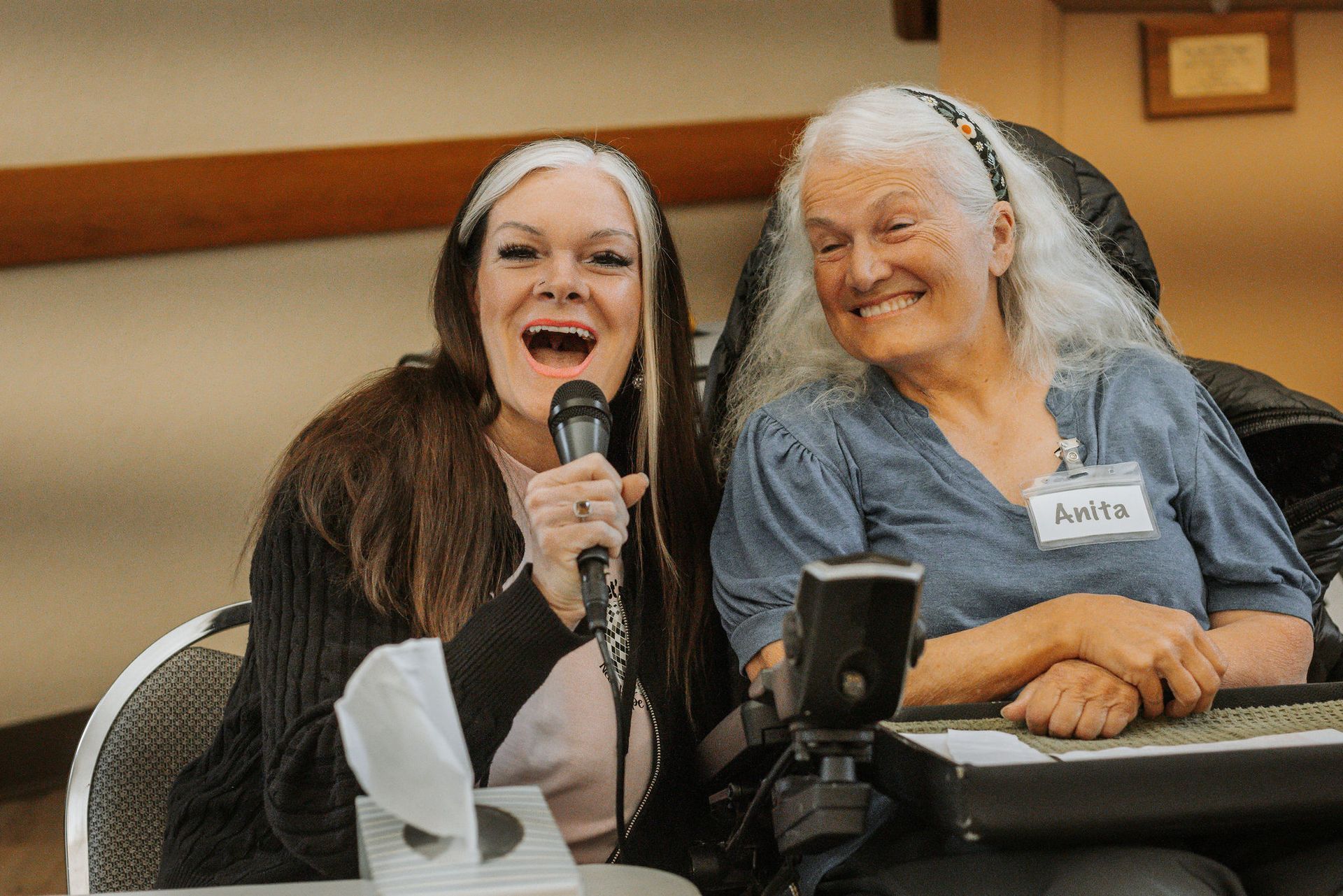 Two women smiling and laughing, one with a microphone, in a room.