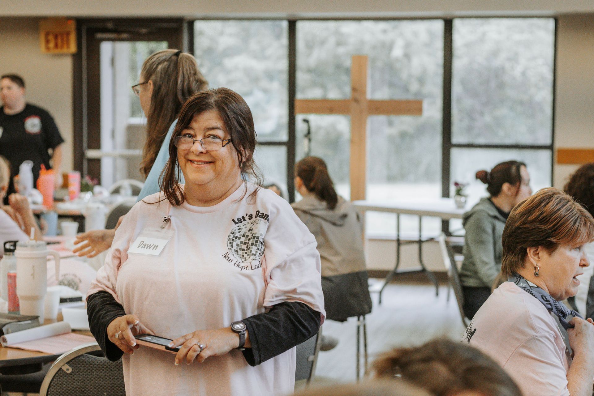 Woman in pink shirt smiles, holding phone. People around tables, cross in background.