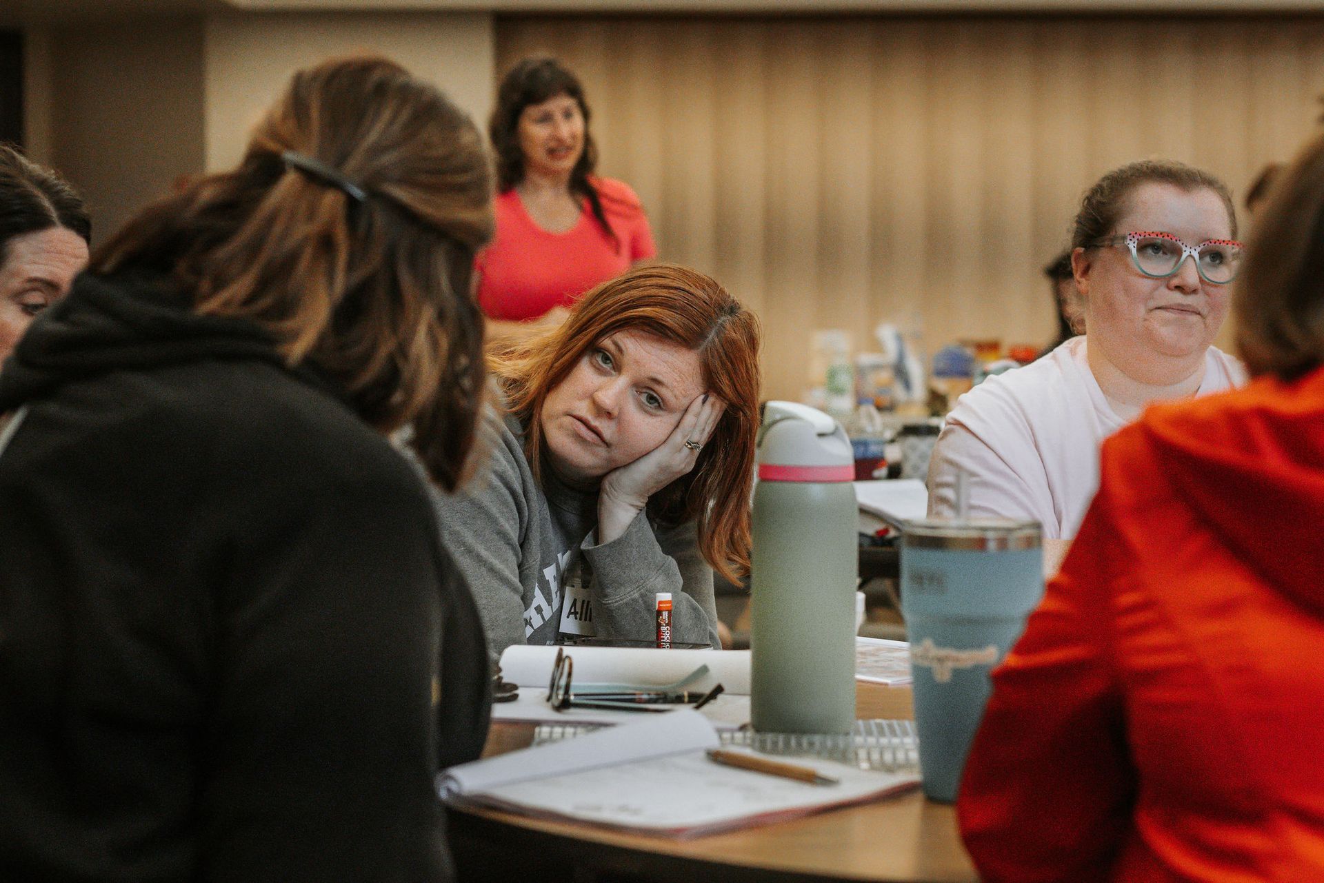 People at a table, some looking at each other. Woman with red hair leans on hand.  A woman wears a red shirt.