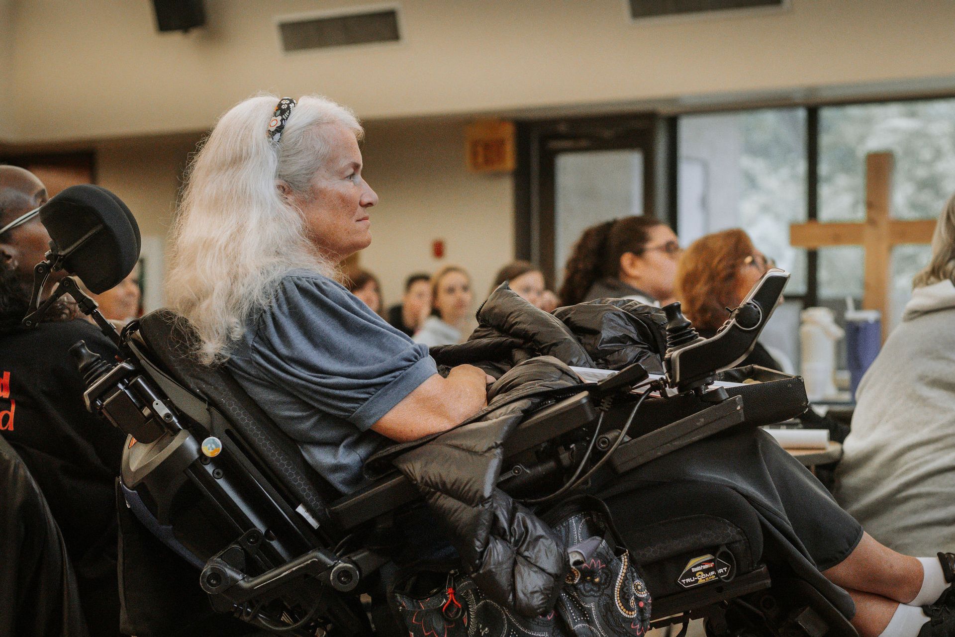 Woman in wheelchair at a meeting; others in attendance in a room. Cross on wall visible.