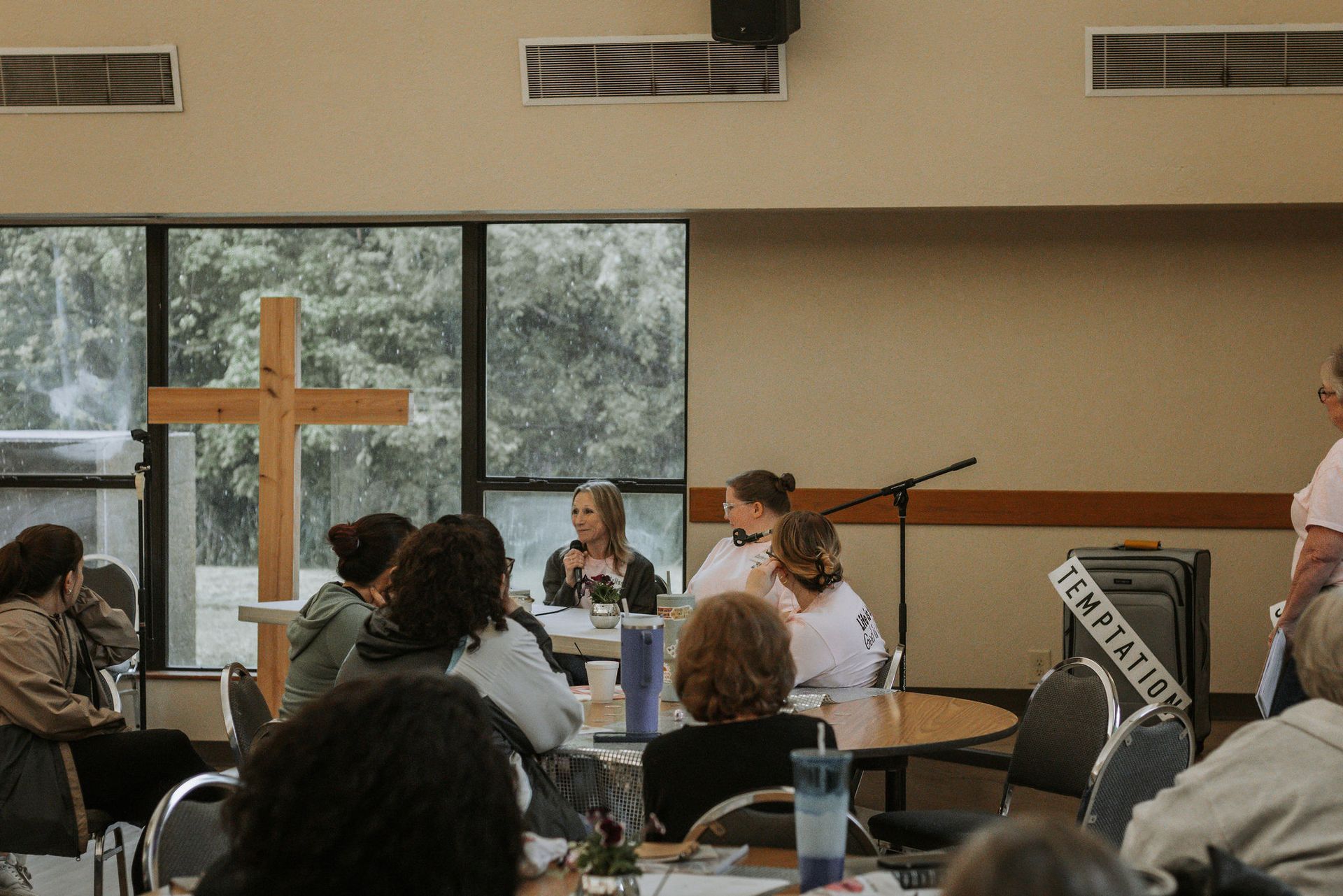 People gathered in a room with a cross, table, and window. Three women speak at a table.