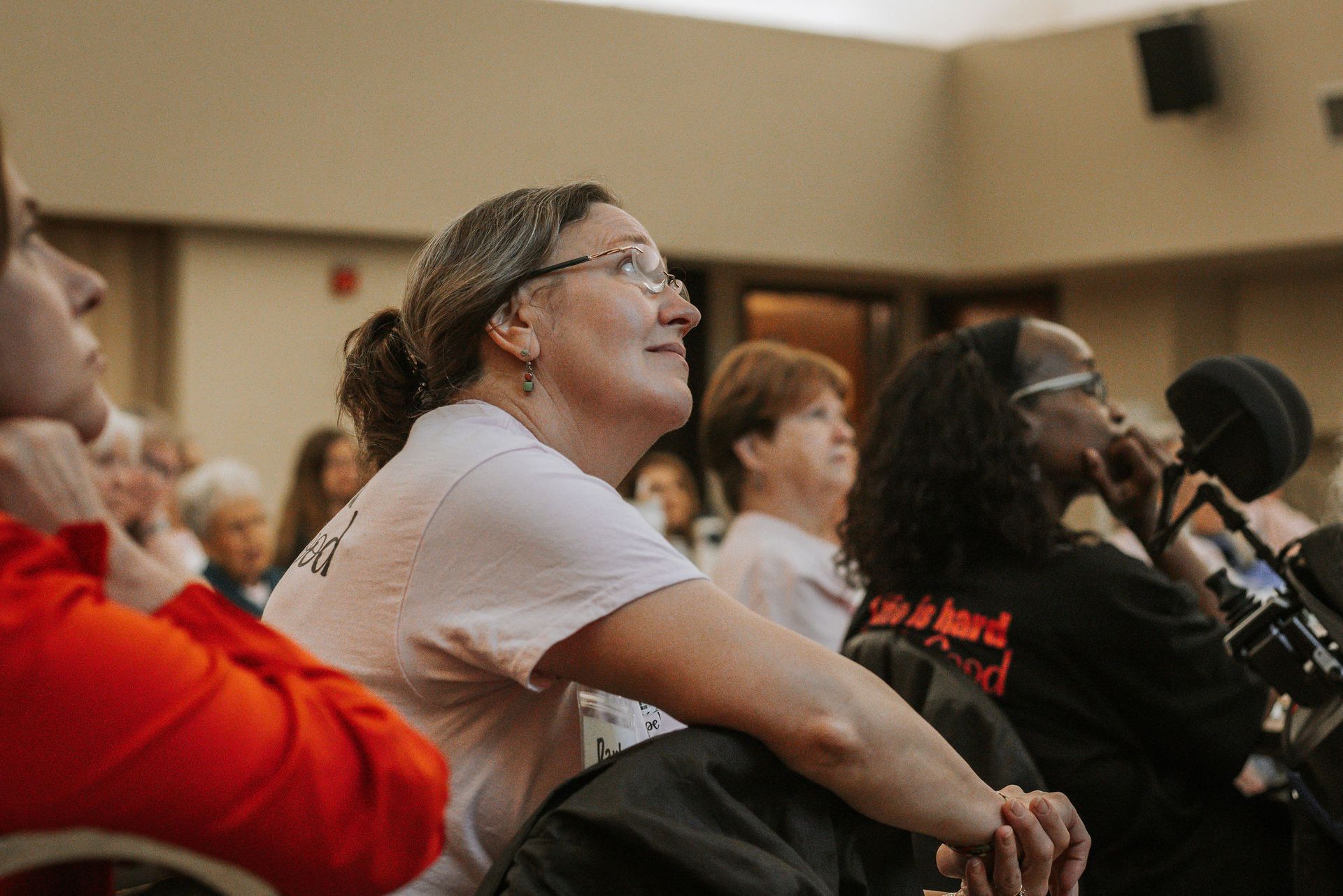 People in a room, listening intently. A woman in glasses and a light shirt looks upward. Others are visible.