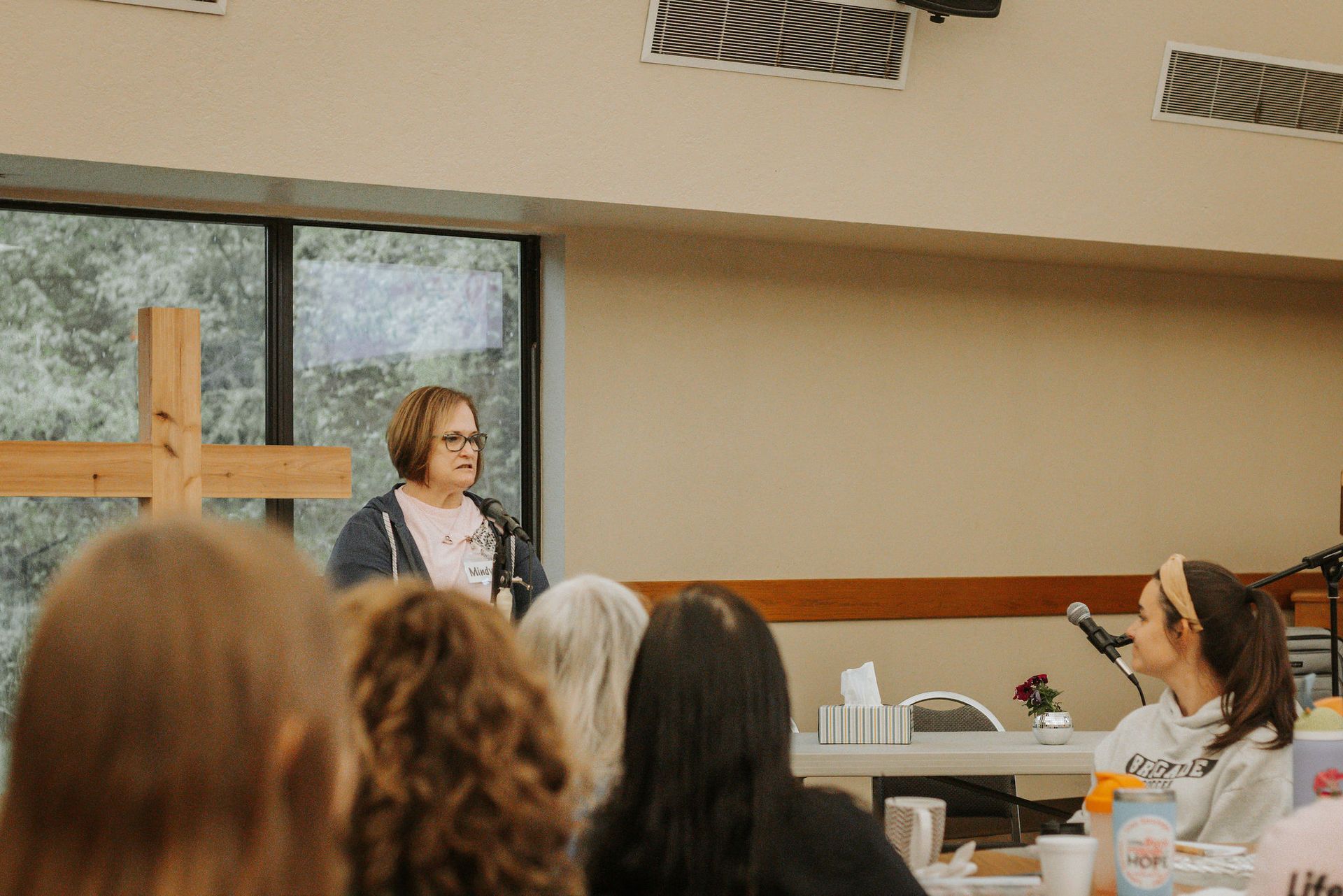 Woman speaking at lectern, facing audience in a room with a wooden cross.