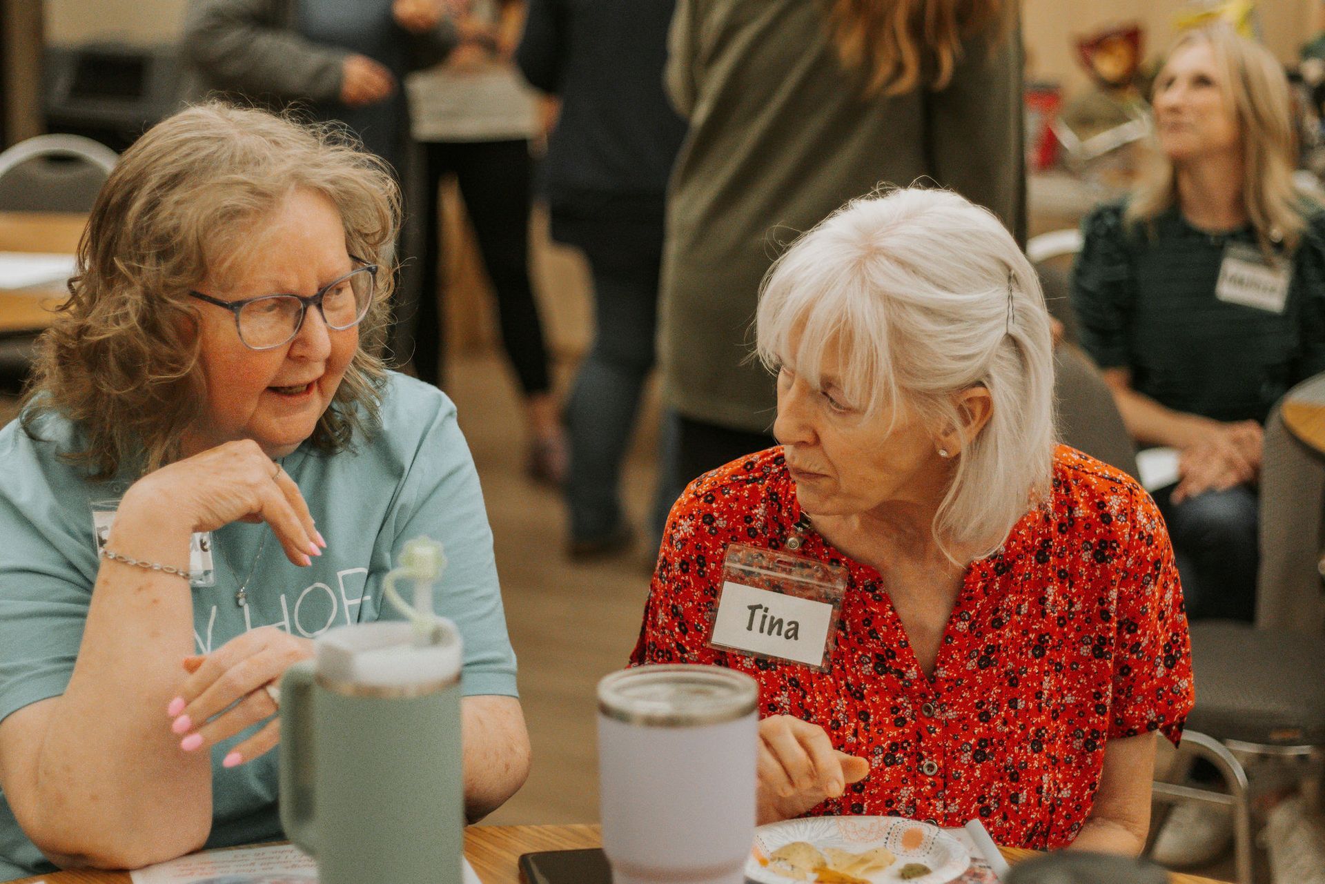 Two women at a table talking. One wears a blue shirt, the other a red patterned shirt. Other people in the background.