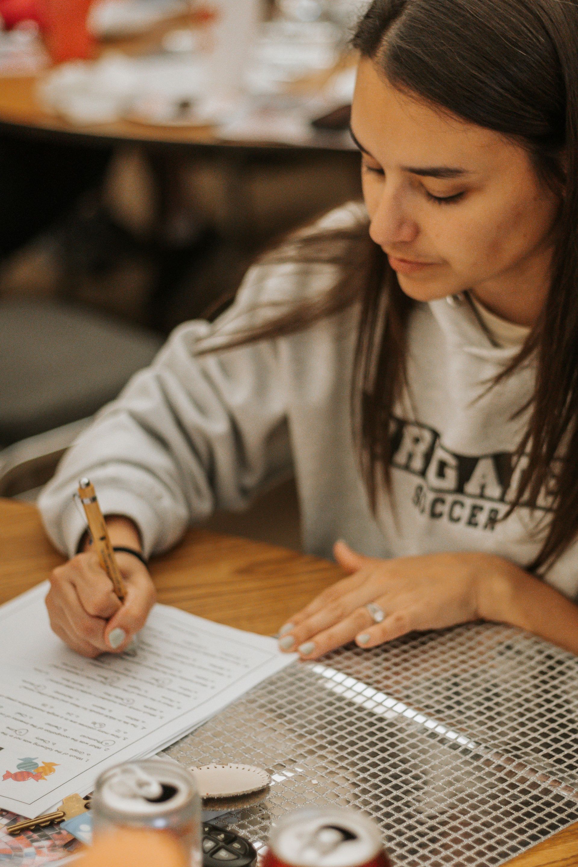 Woman in a gray sweatshirt writing at a table, silver mat, soda cans, and wood surface.