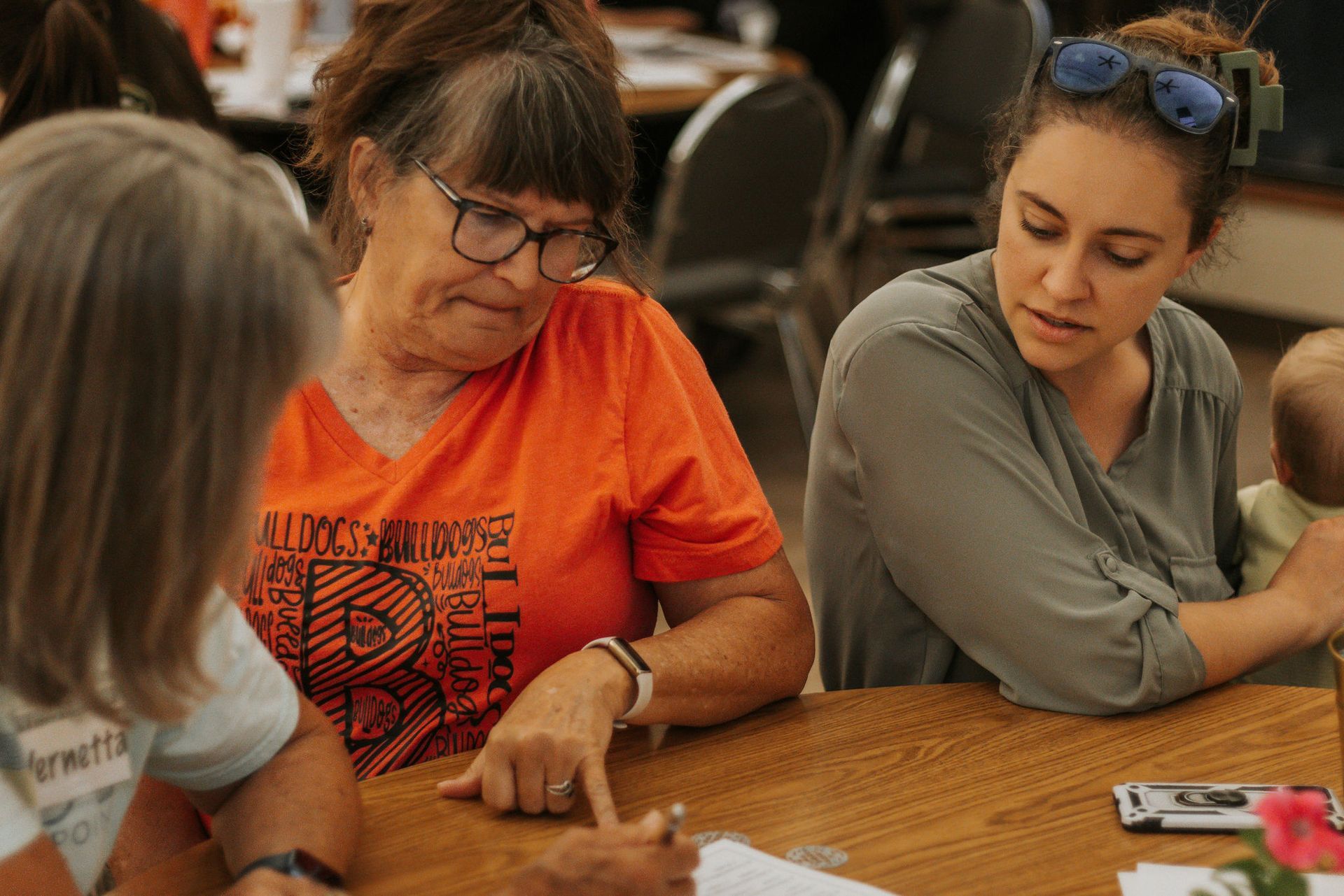 Three women at a table; one points to something while others watch. Sunlight, wood table, bright colors.