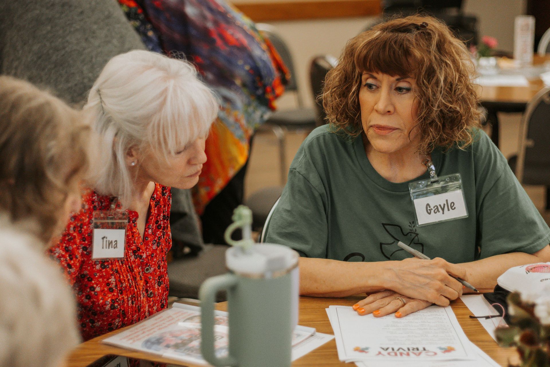 Two women with name tags seated at a table, looking at papers. One wears a green shirt, one a red shirt.