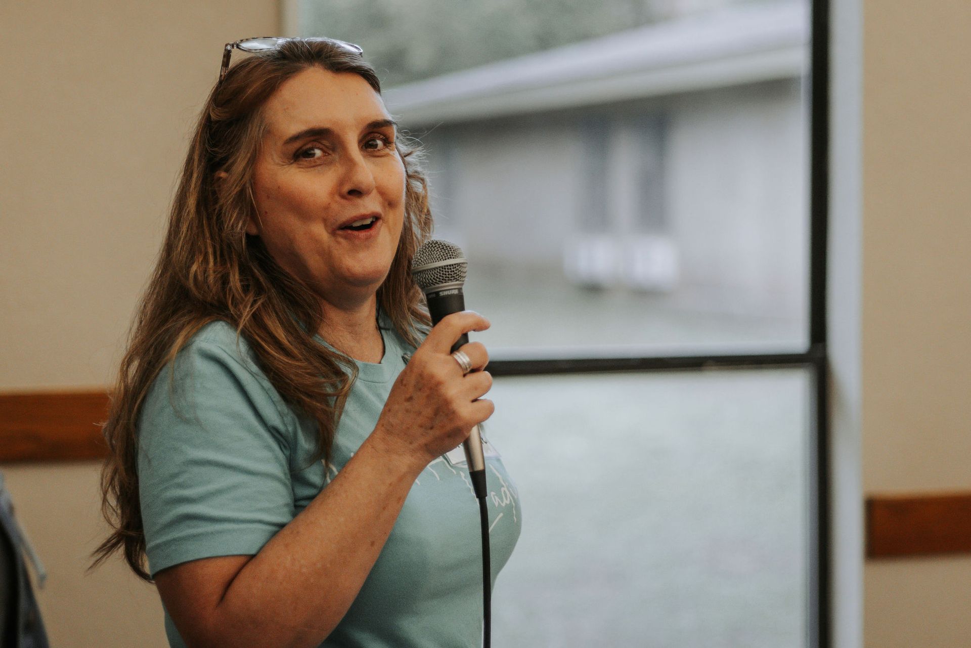 Woman with long brown hair speaks into a microphone, wearing a blue t-shirt, standing indoors near a window.