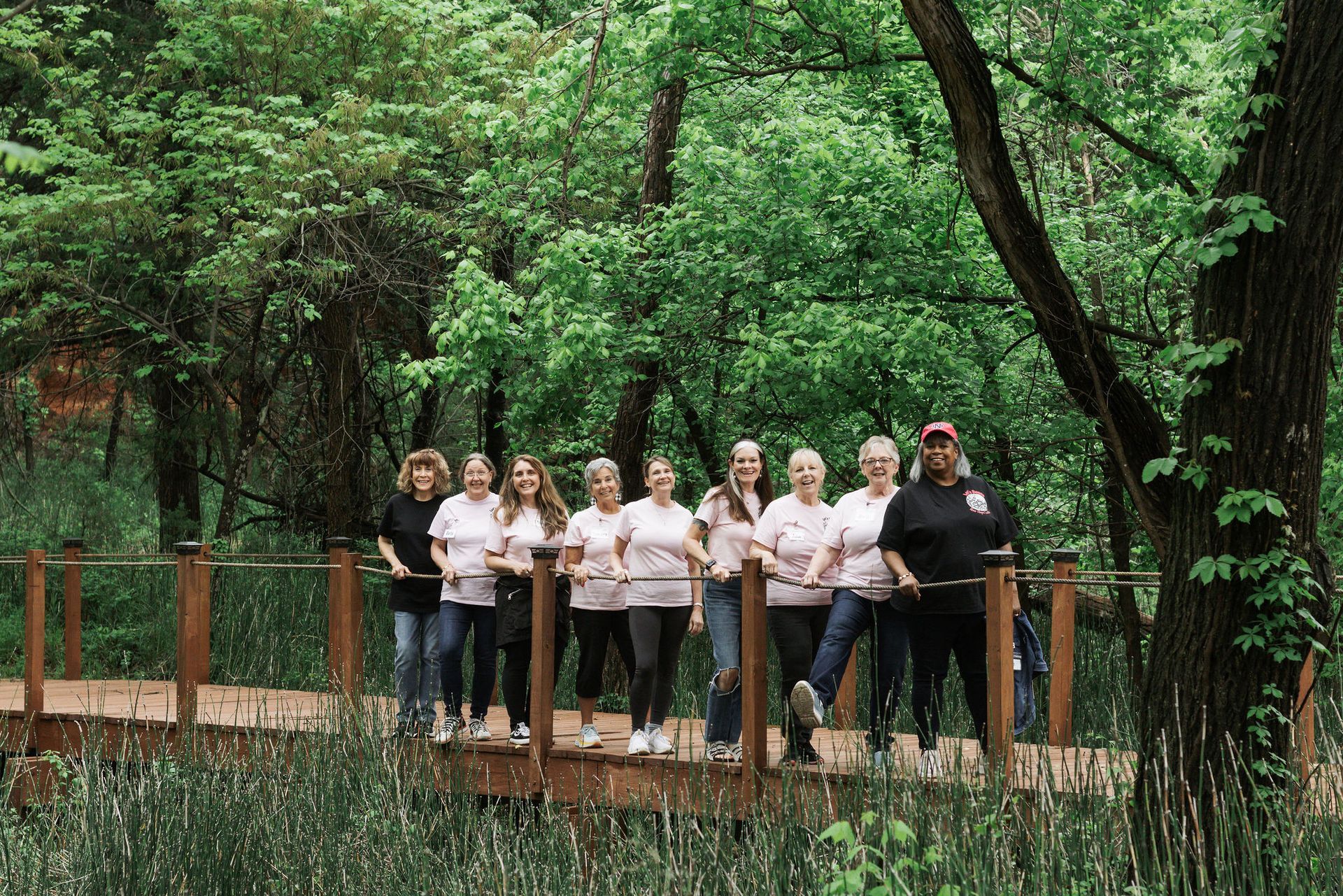 Group of women on a wooden walkway in a forest, smiling at the camera. Most wear pink shirts and jeans.