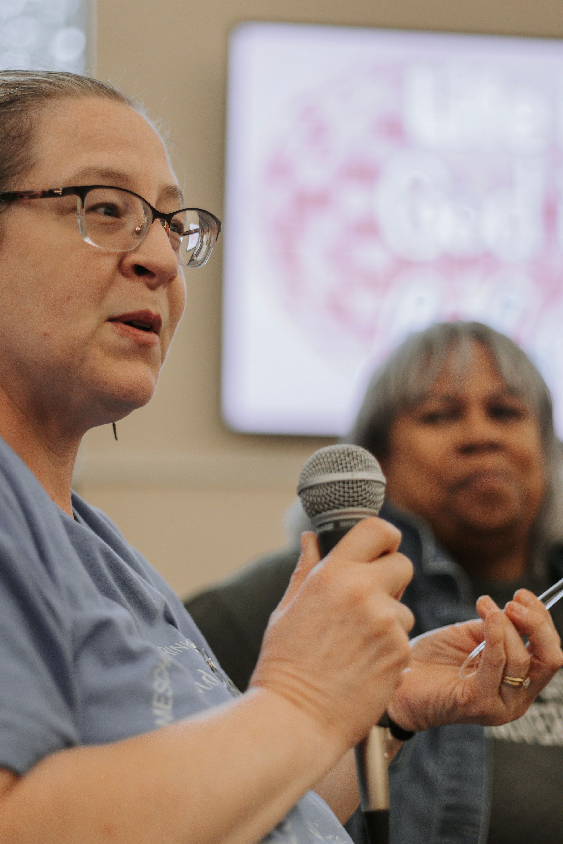 Woman with glasses speaking into a microphone. Another woman is seated in the background.