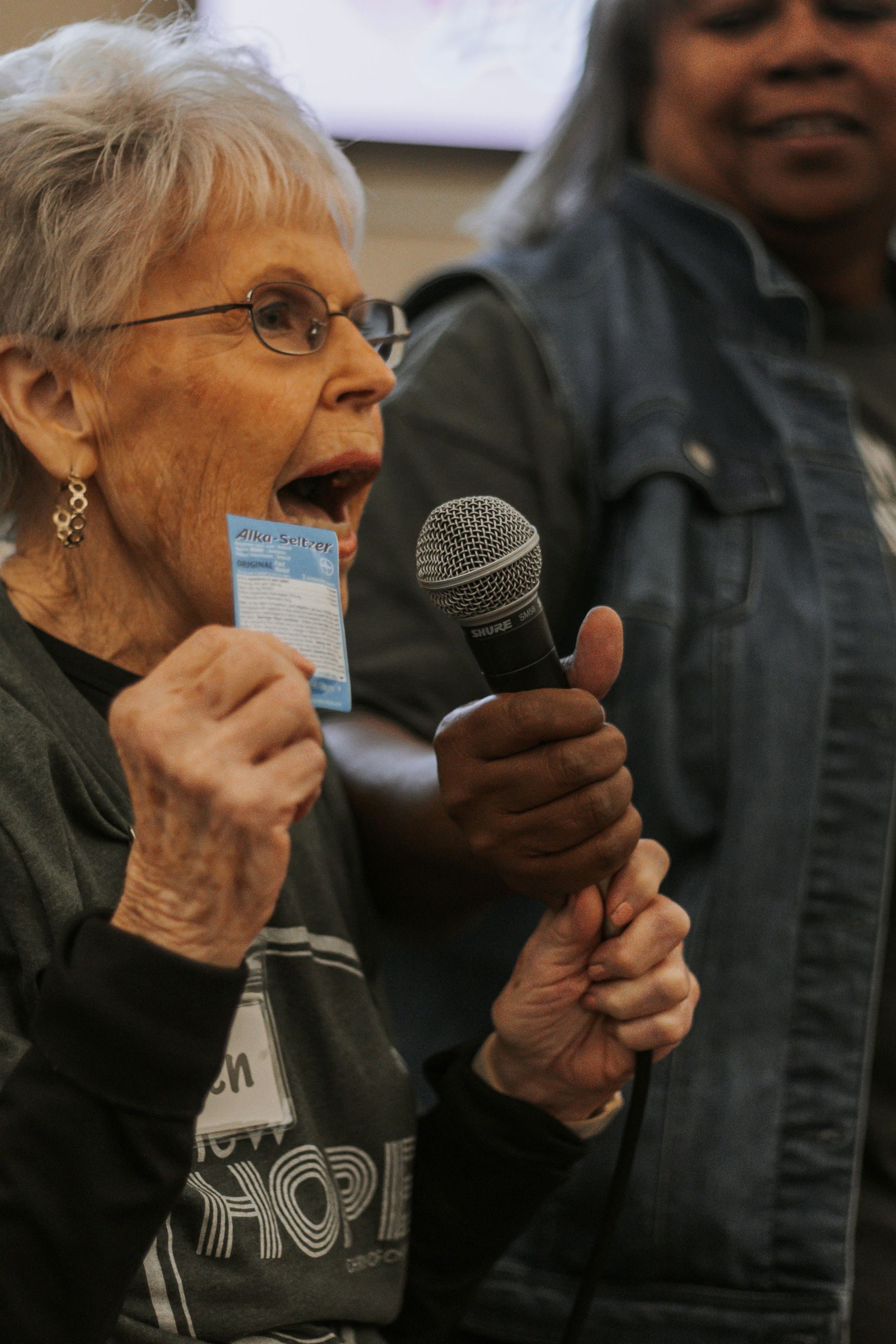 Excited senior woman holding ticket, speaking into microphone. Another woman stands nearby. Indoor setting.