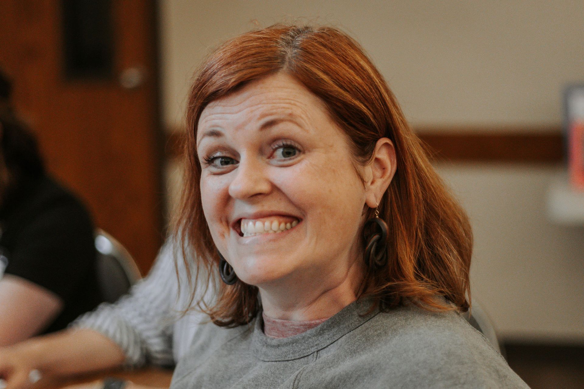 Woman with red hair smiles widely, indoor setting, wearing gray sweater.