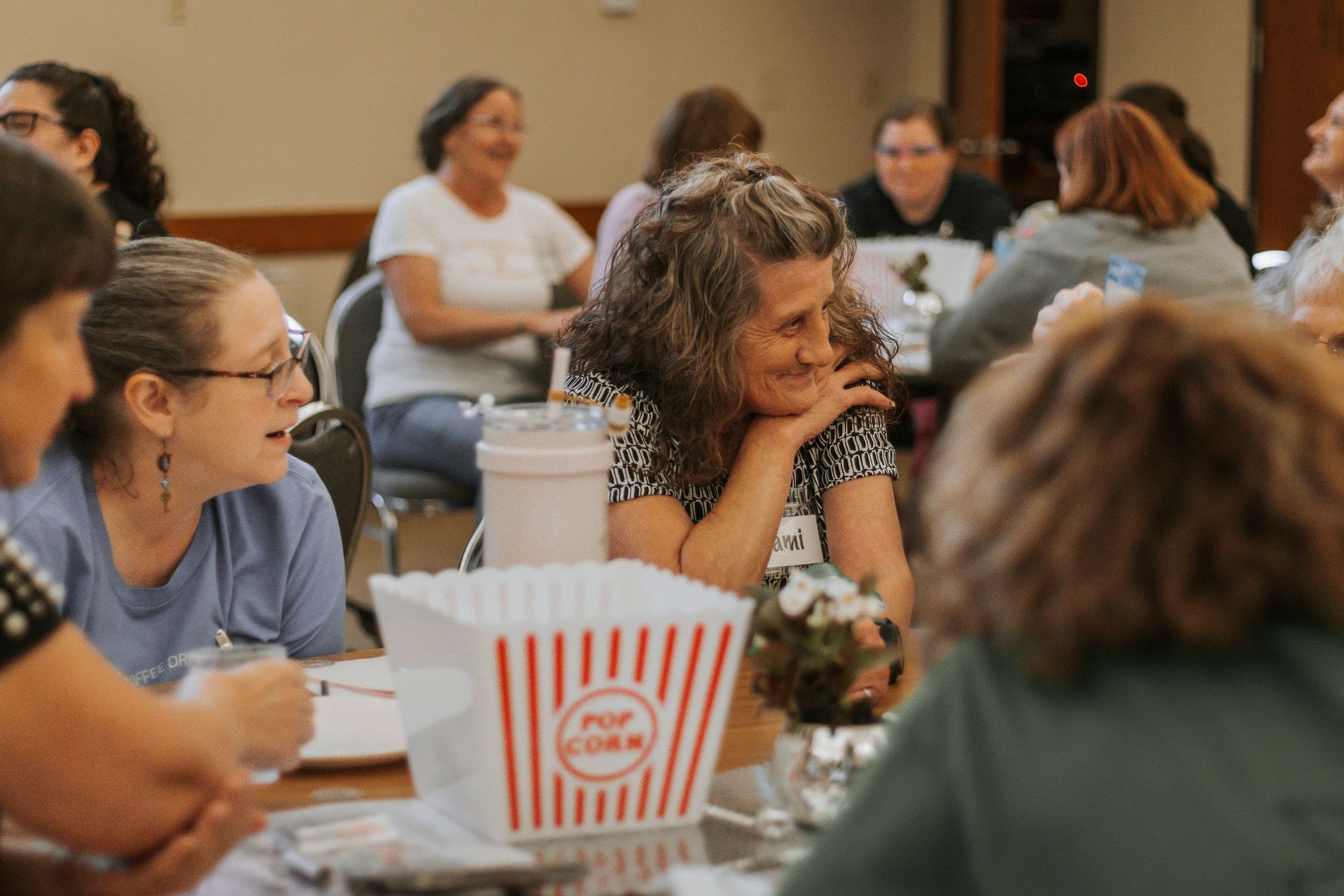 People gathered around tables, smiling, in a room with a popcorn bucket, flowers, and a neutral background.