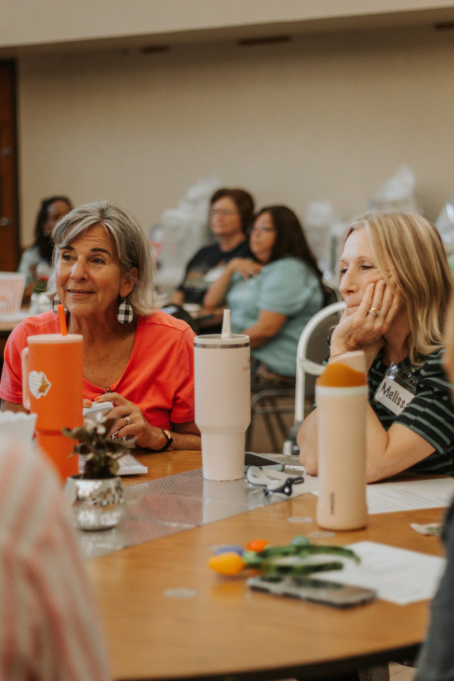 Attendees at a table listen intently. Two women in the foreground, one in red, one in stripes, smiling.