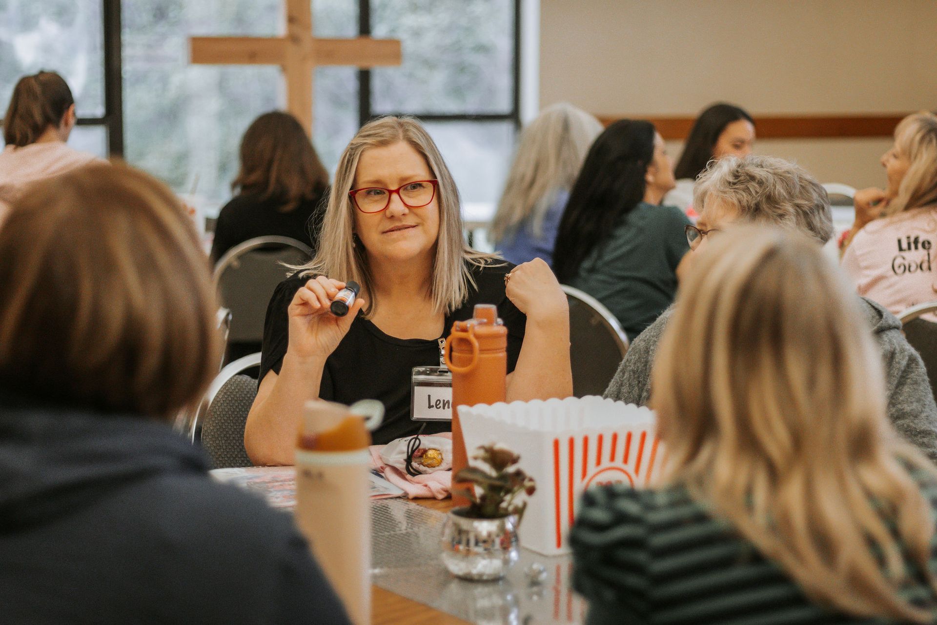 Woman smiling at table with others in a room; cross in background; popcorn.
