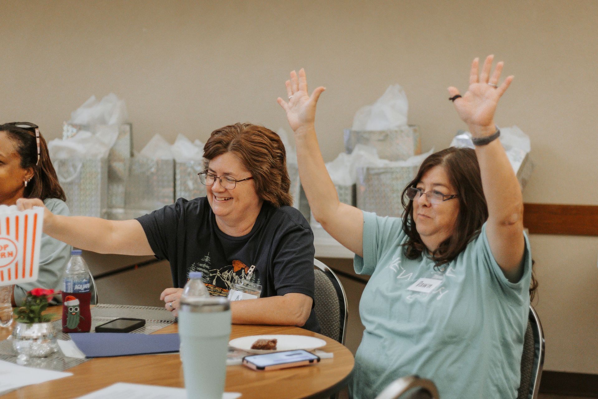 Two women at a table raise their hands, smiling. One holds popcorn. White gift bags in the background.