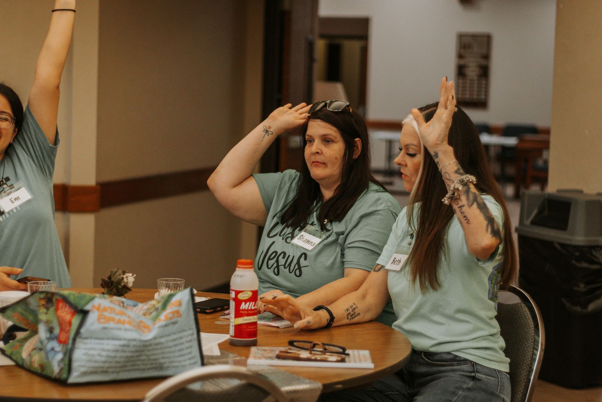 Three women in teal shirts with arms raised, seated at a round table, indoors.