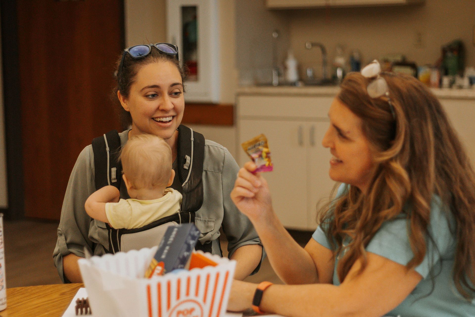 Woman shows a snack to a baby in a carrier; another woman smiles, sitting at a table in a bright room.