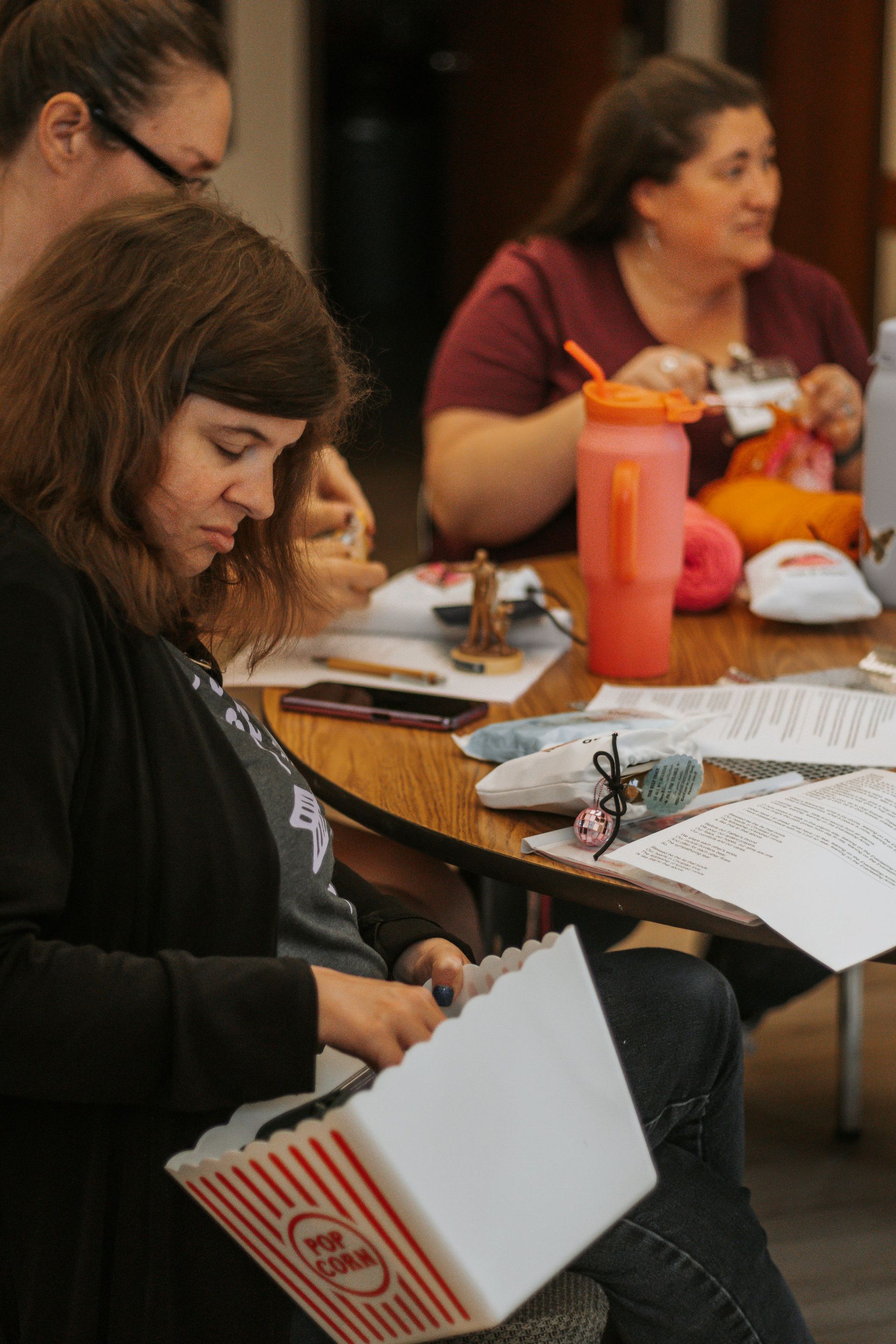 Woman in a dark sweater eats popcorn at a table with other women. They appear to be knitting.