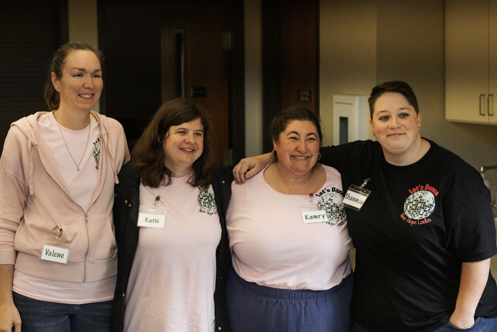 Four women smiling, wearing pink and black shirts, arm-in-arm in a brightly lit room.