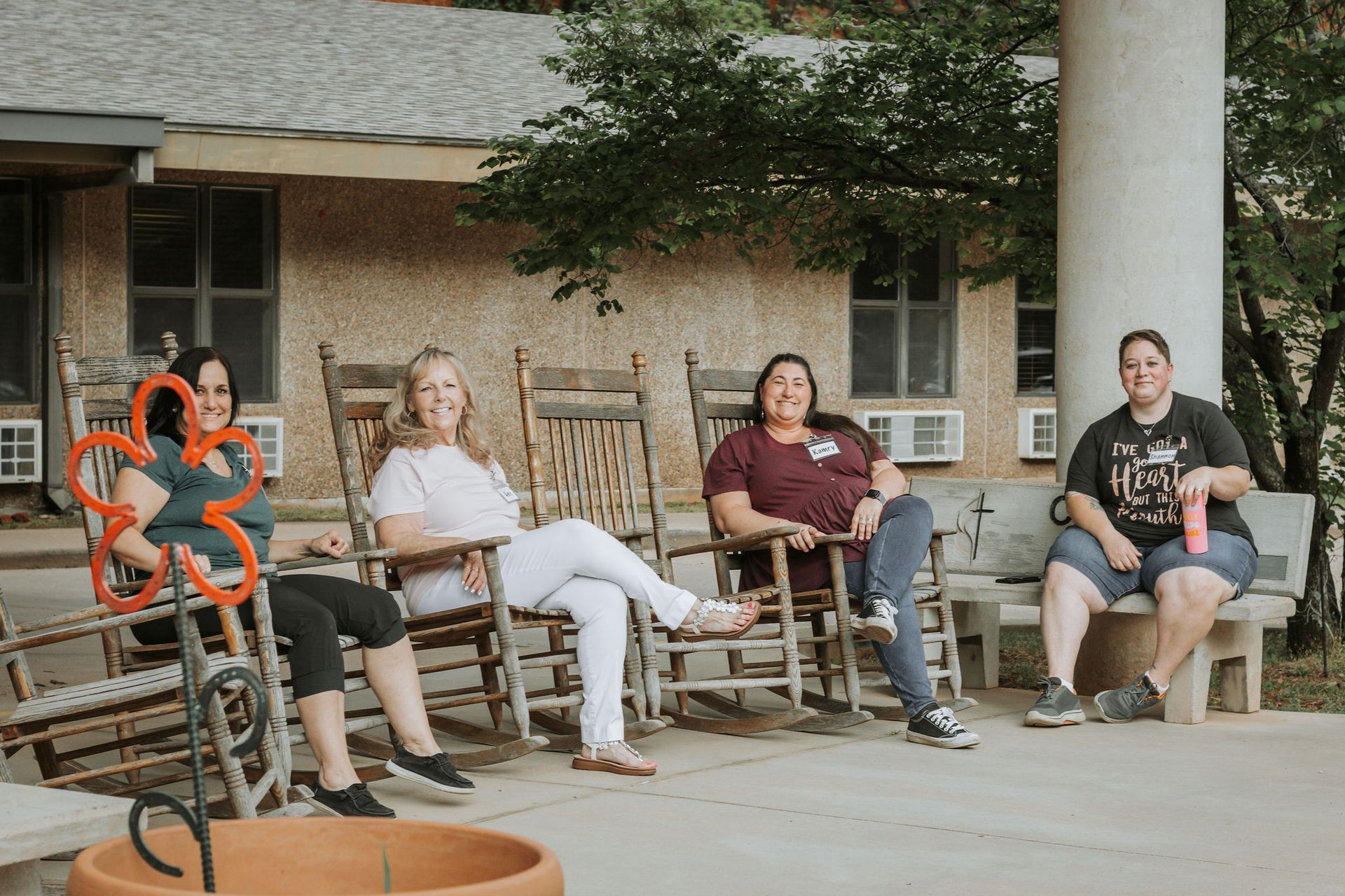 Four women in rocking chairs on a porch. One holds a flower decoration. Exterior setting.