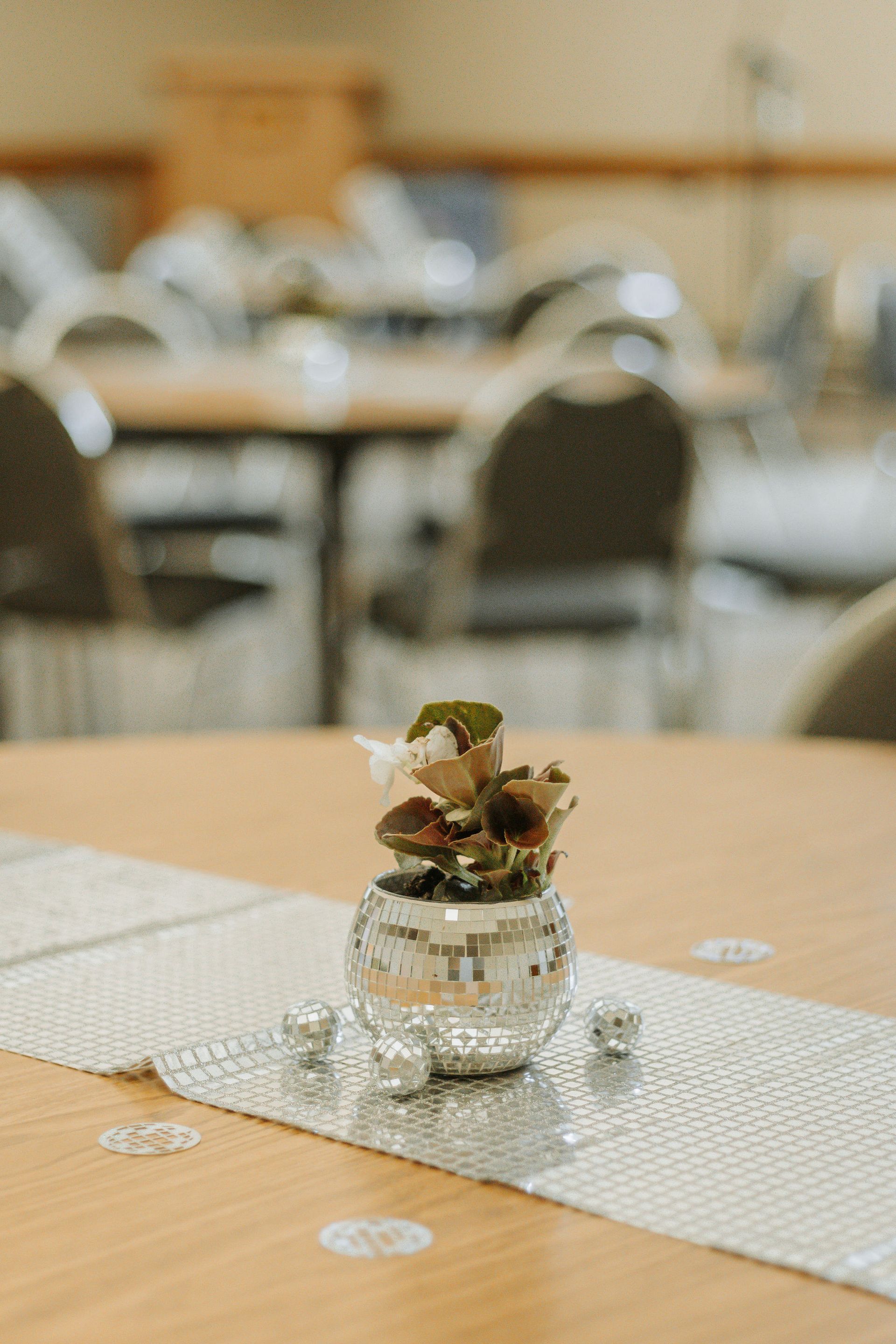 A table with a mirrored vase of flowers on a silver runner, set in a room with tables and chairs.
