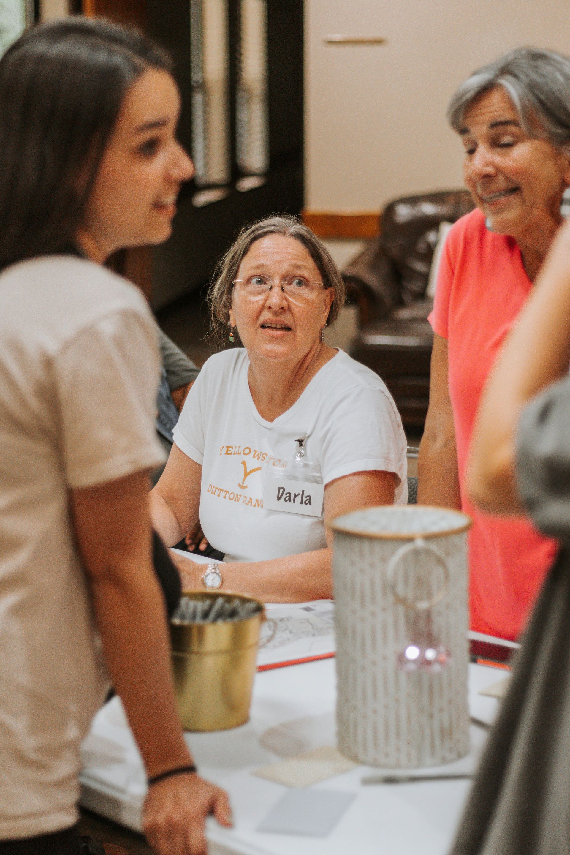 Three women at a table. Woman in the middle with glasses looks up, others smile. Indoor setting.