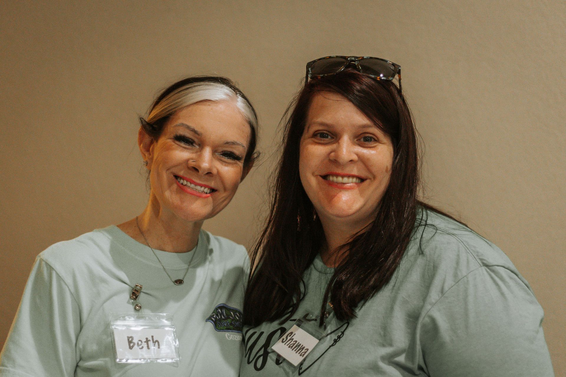 Two smiling women wearing teal shirts, name tags, and sunglasses on the head, stand side-by-side.