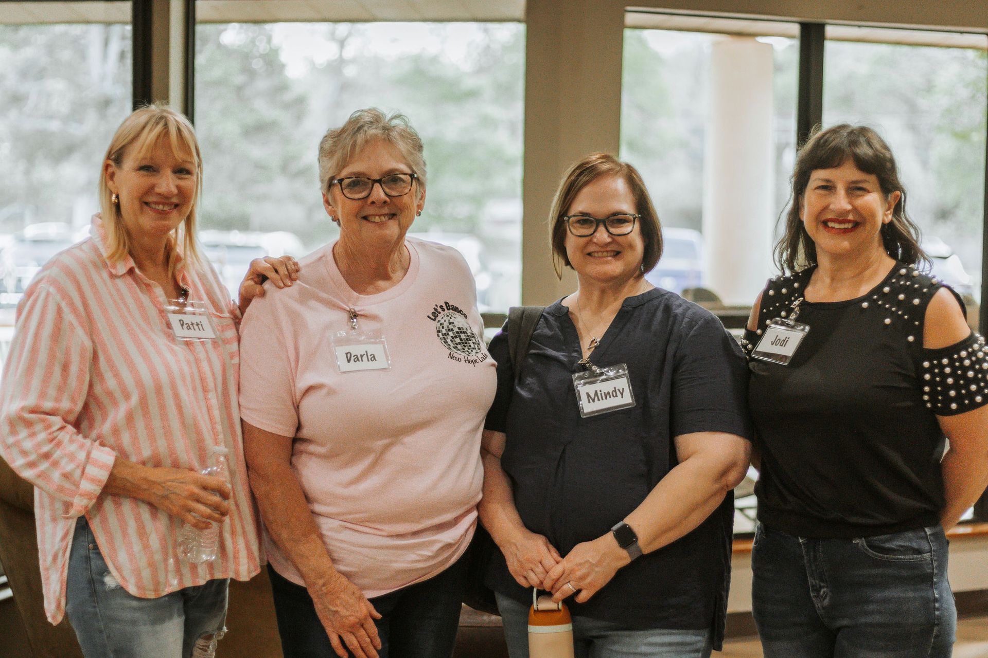 Four smiling women pose together indoors; they wear casual clothing and name tags.