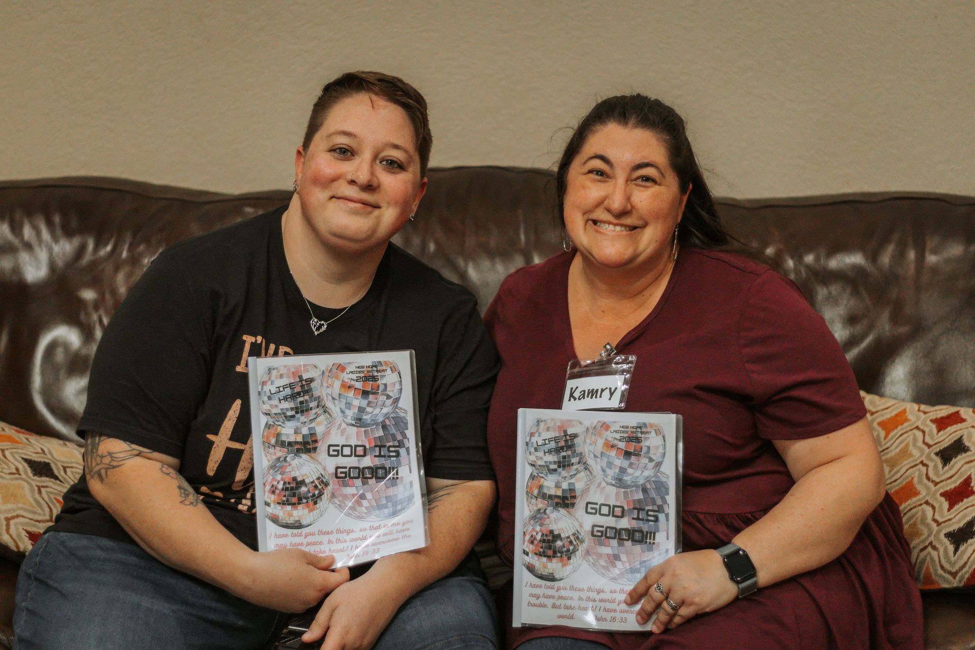 Two women on a couch holding books, smiling. One wears black, the other burgundy.