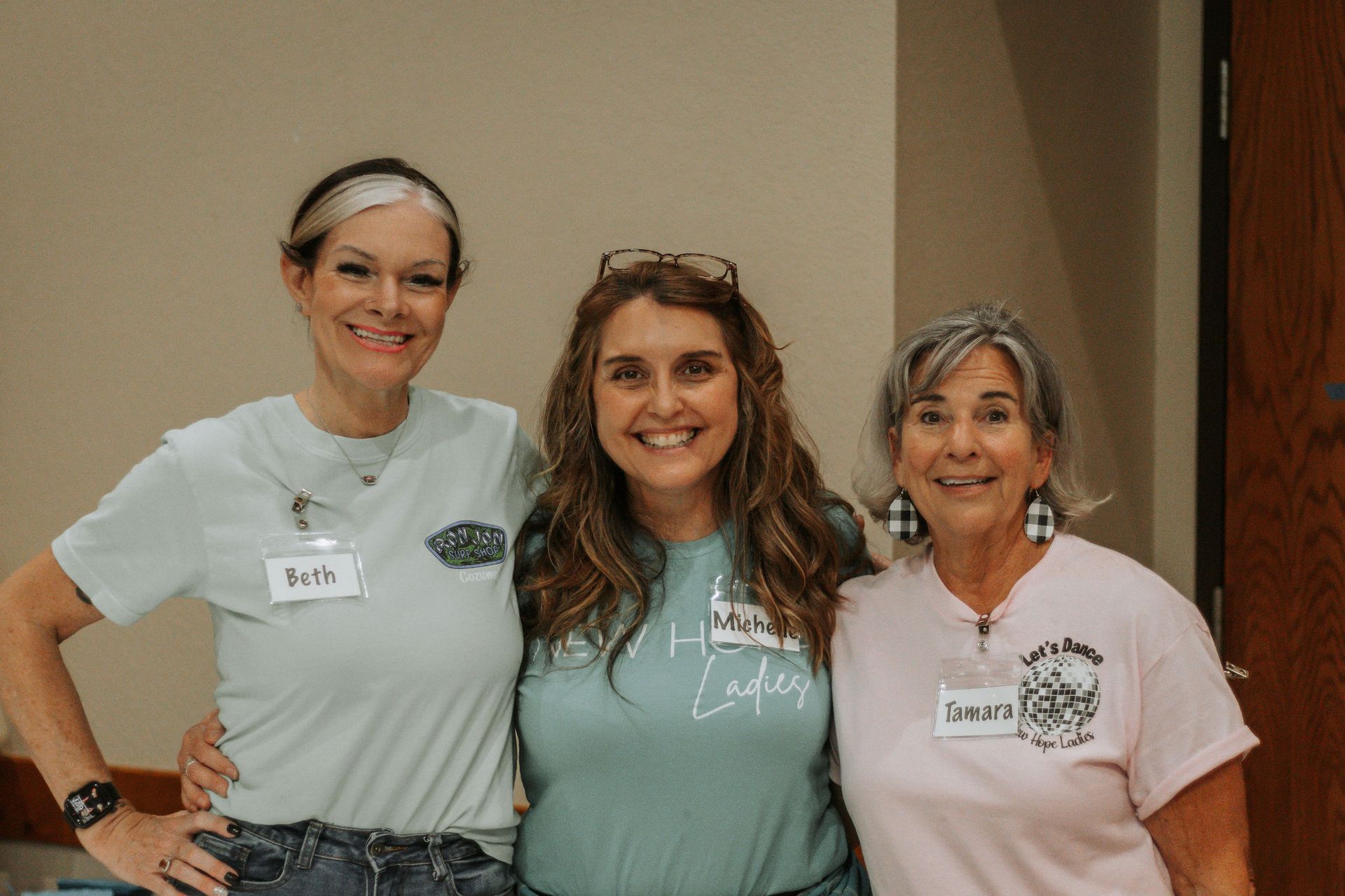 Three women smiling, hugging; indoors, light-colored walls. Middle woman wears glasses, others have name tags.