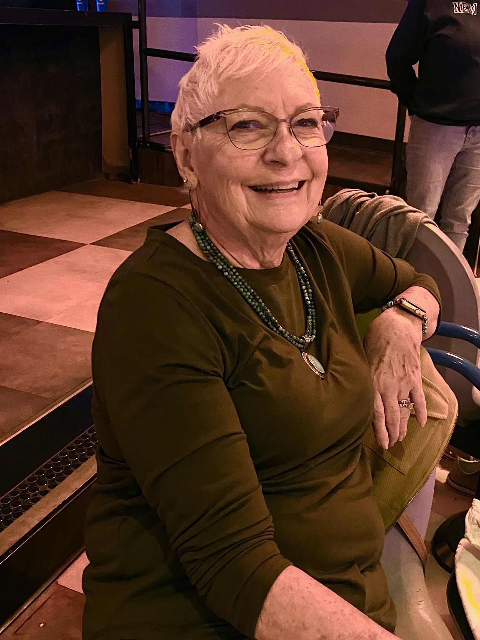 An elderly woman wearing glasses and a green shirt is sitting at a table.
