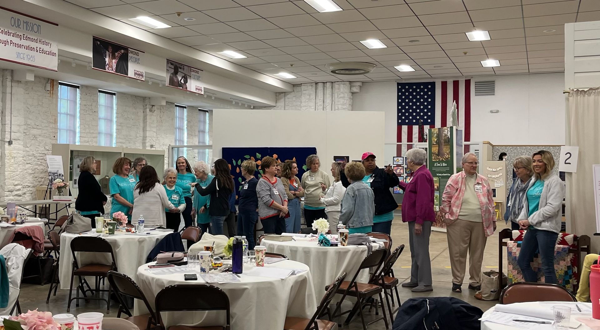 A group of people are standing around tables in a room with an american flag on the wall.