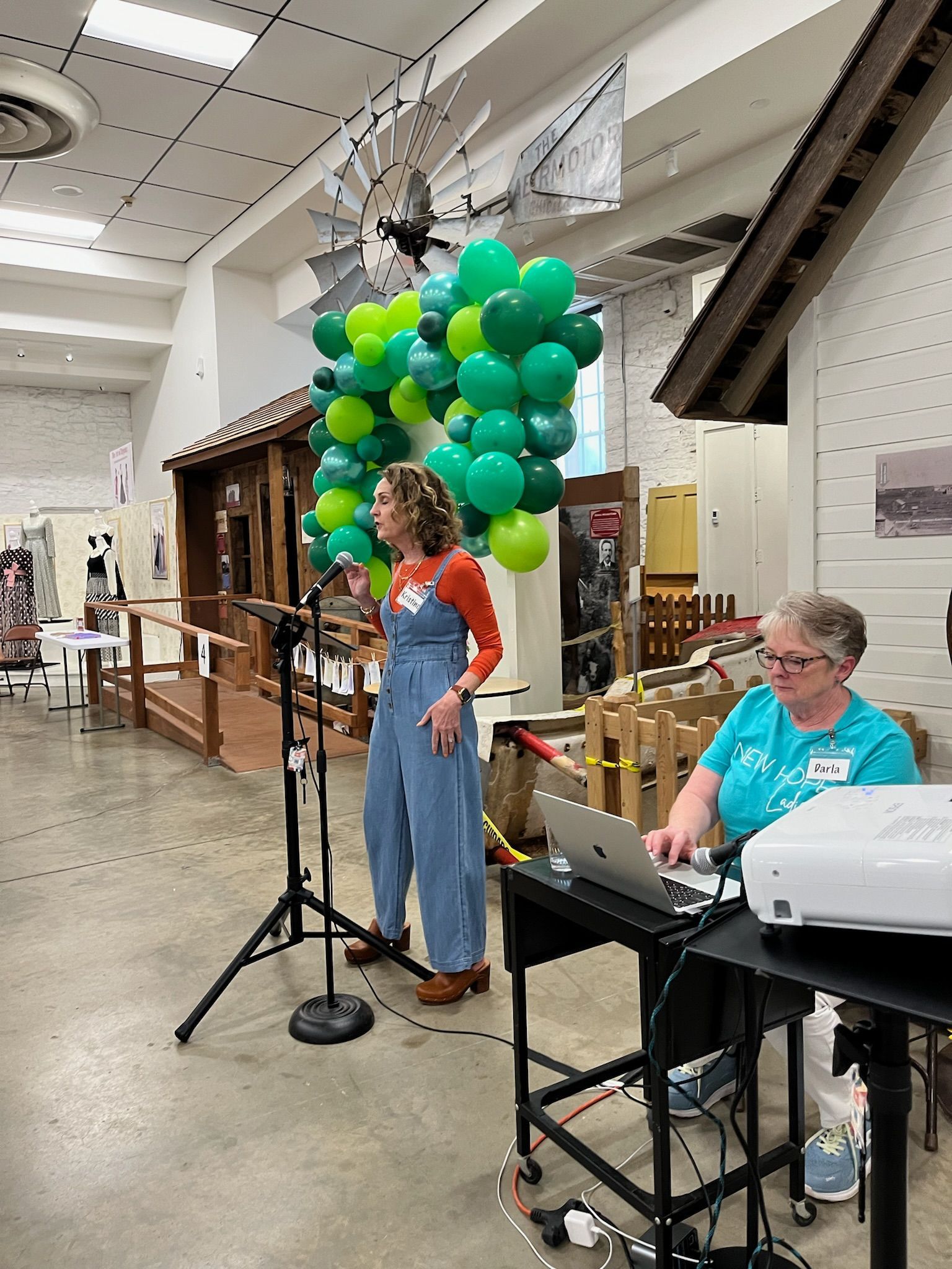 Two women are standing in front of a microphone in a room with green and yellow balloons.