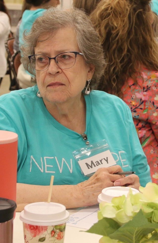 An older woman wearing glasses and a name tag is sitting at a table with a cup of coffee.