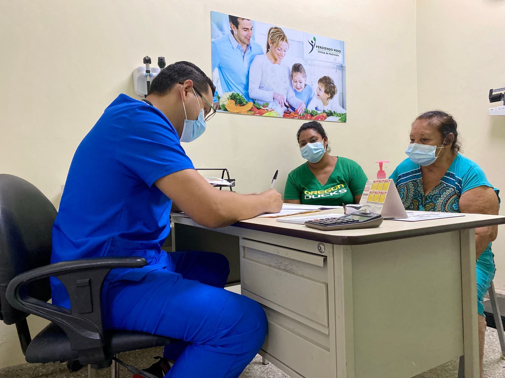A doctor is sitting at a desk talking to two women wearing masks.