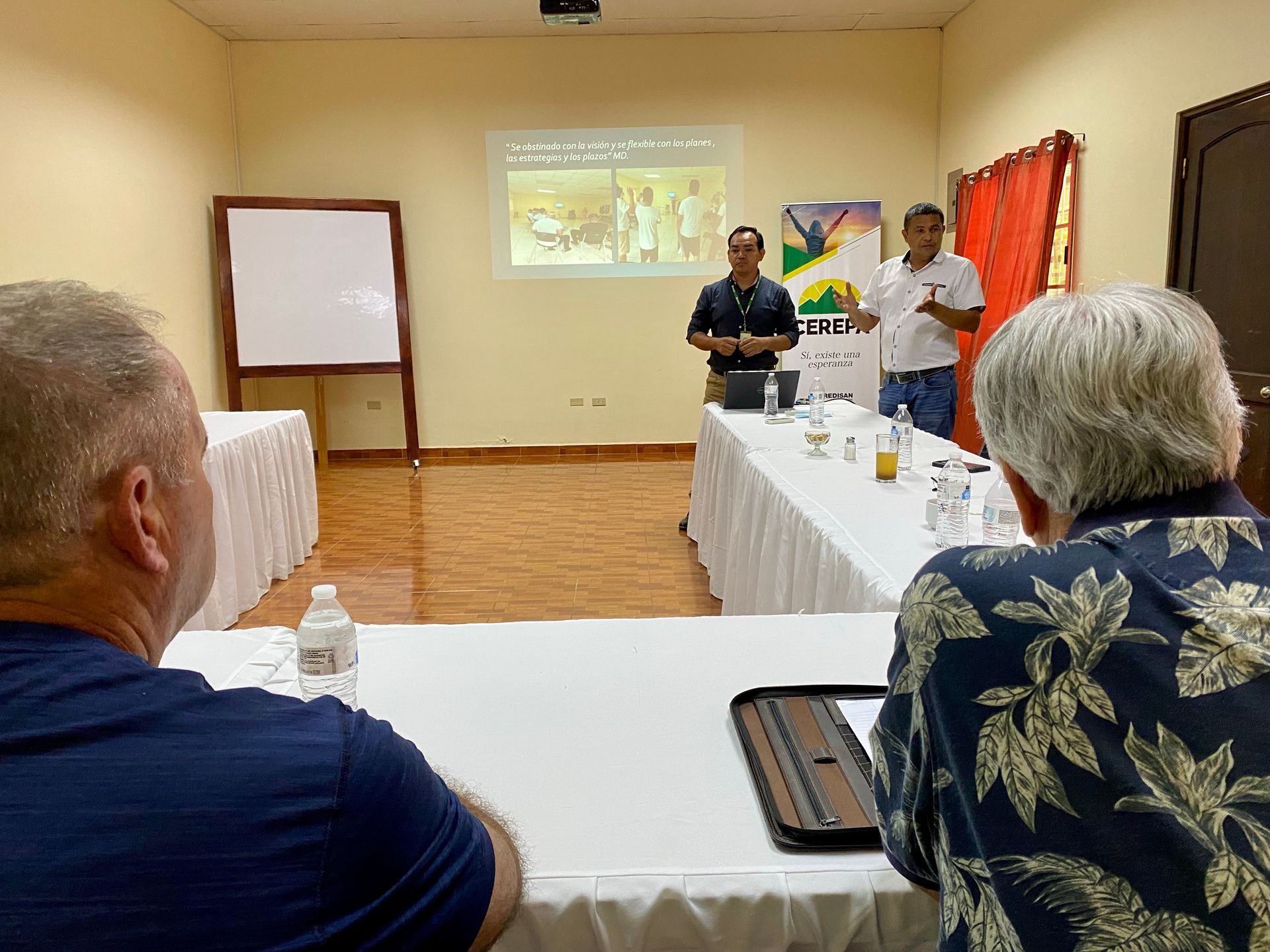 A group of men are sitting at tables in a conference room watching a presentation.