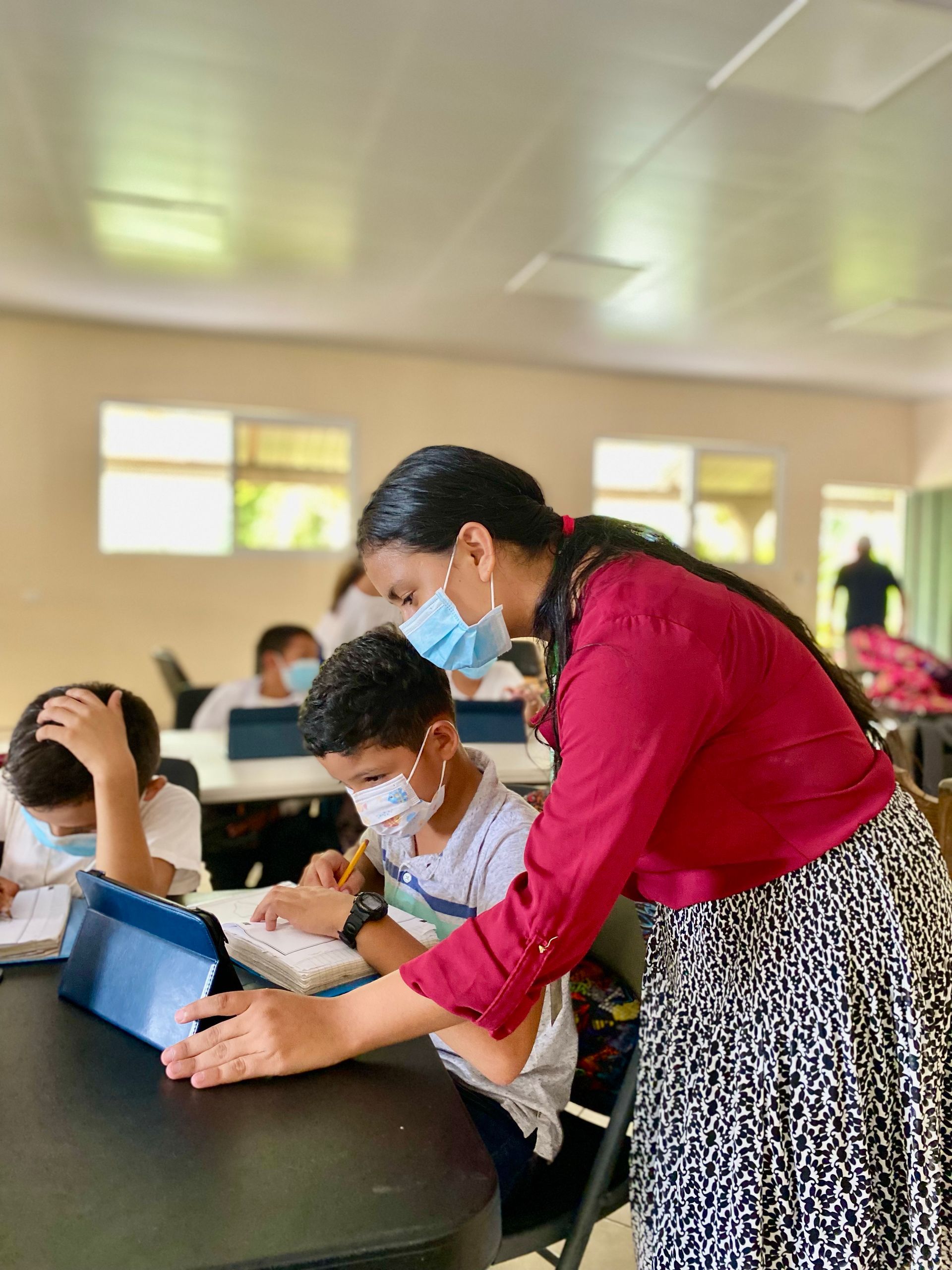 A woman wearing a mask is helping a boy with his tablet in a classroom.