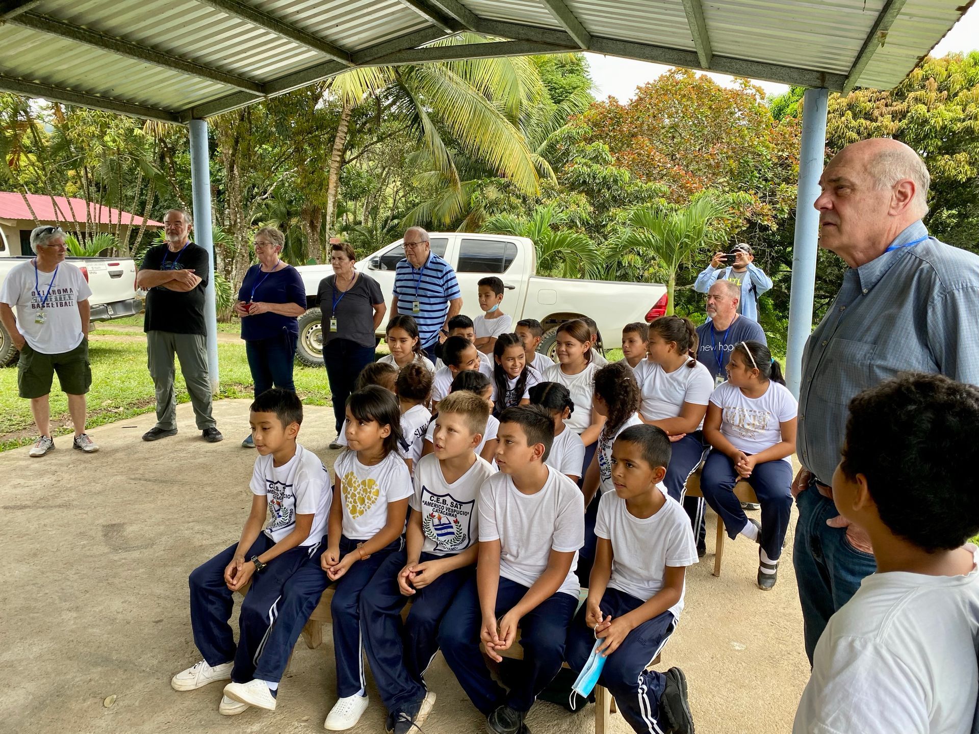 A group of children are sitting under a canopy while a man talks to them.